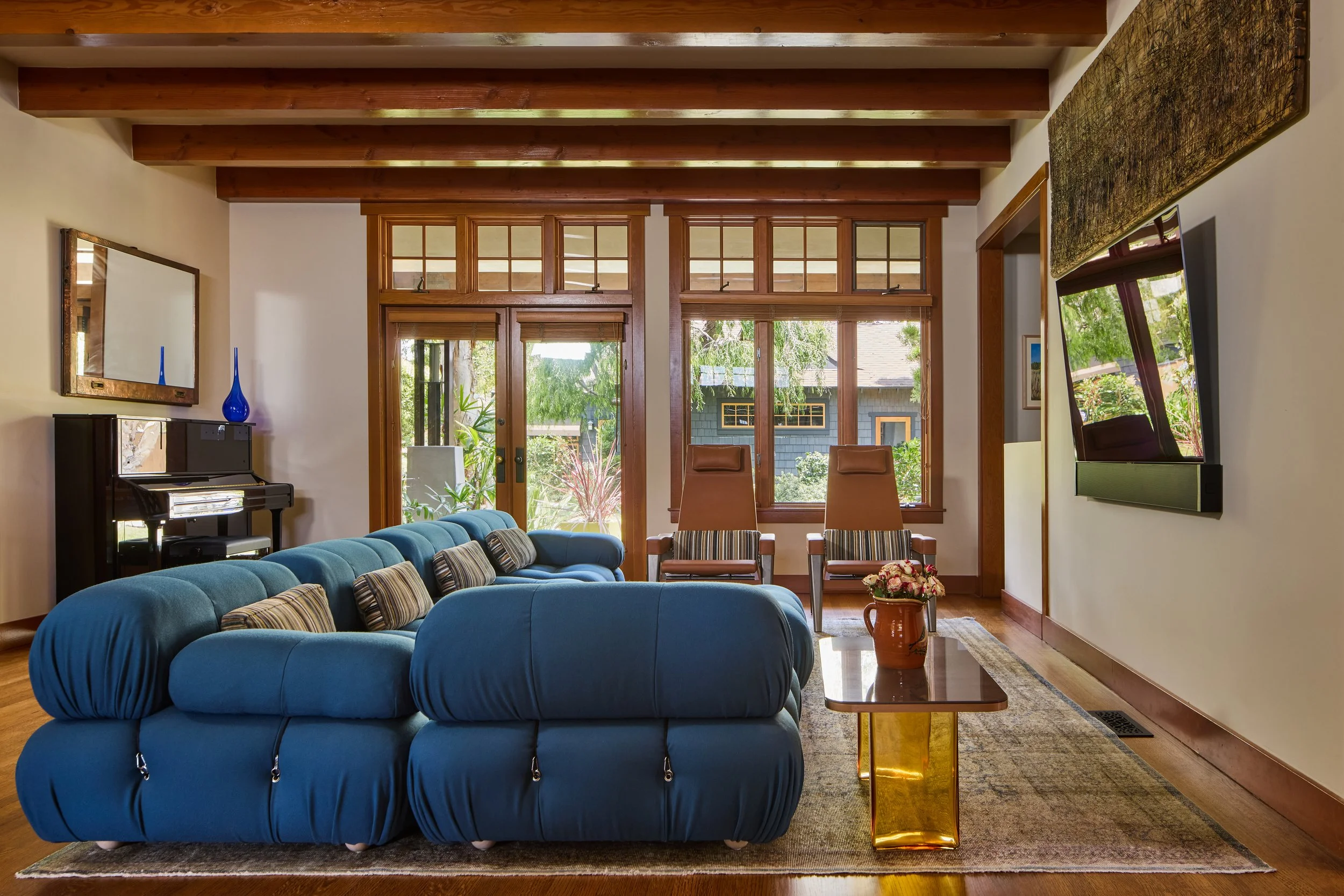 This beautifully designed living room showcases a harmonious blend of modern and craftsman architectural styles. The space features exposed wooden ceiling beams, warm hardwood floors, and large grid windows that flood the room with natural light. A p