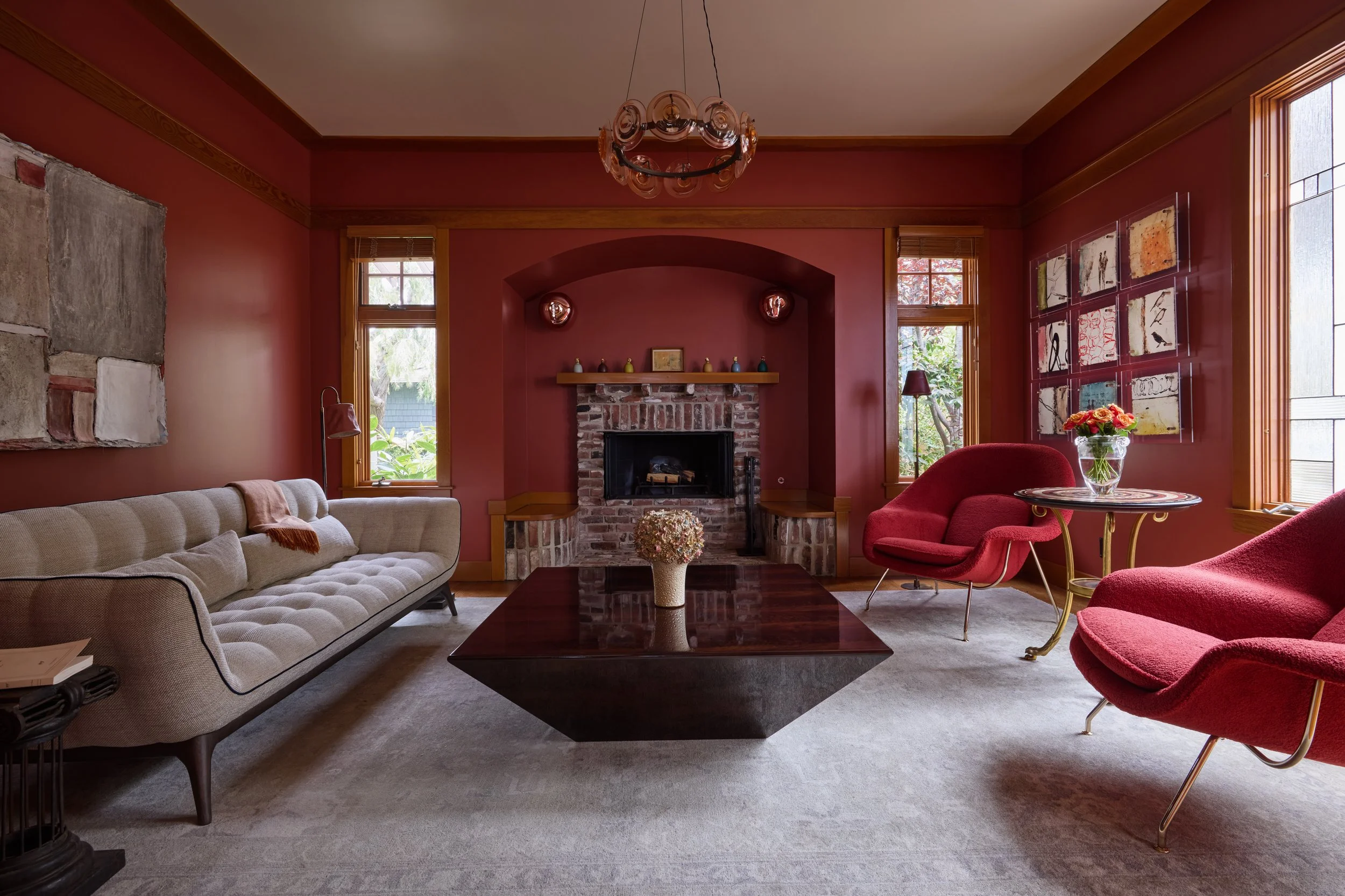 This sophisticated living room captures the perfect fusion of vintage charm and modern design. Rich, deep red walls create a bold backdrop, complemented by warm wood trim and a rustic exposed brick fireplace at the center. A tufted, mid-century moder