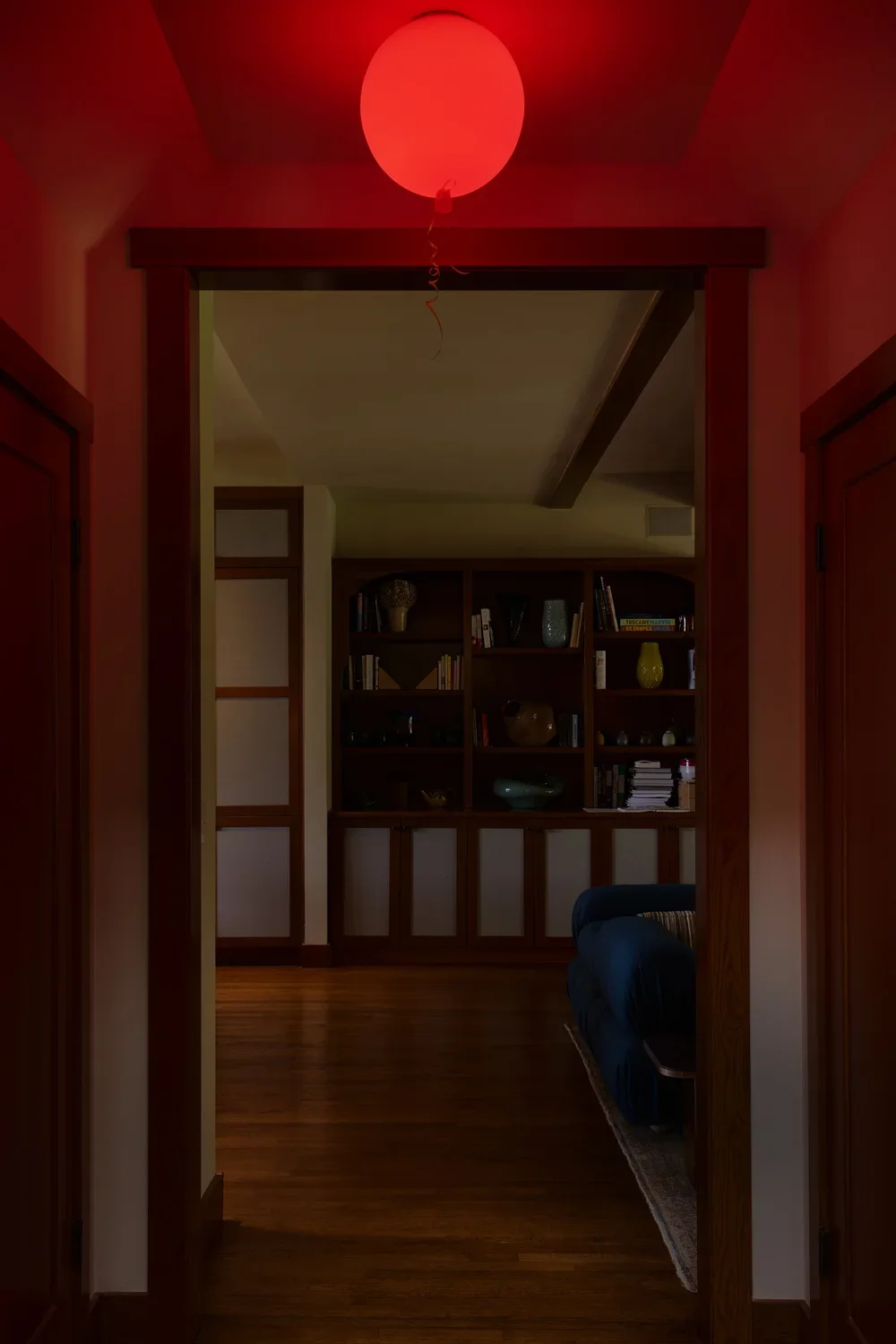 A dramatic hallway view in a Pacific Palisades home, featuring a striking red orb ceiling light that casts a warm glow. The passage opens into a cozy living space with custom wood cabinetry, built-in bookshelves, and curated decorative objects. A dee