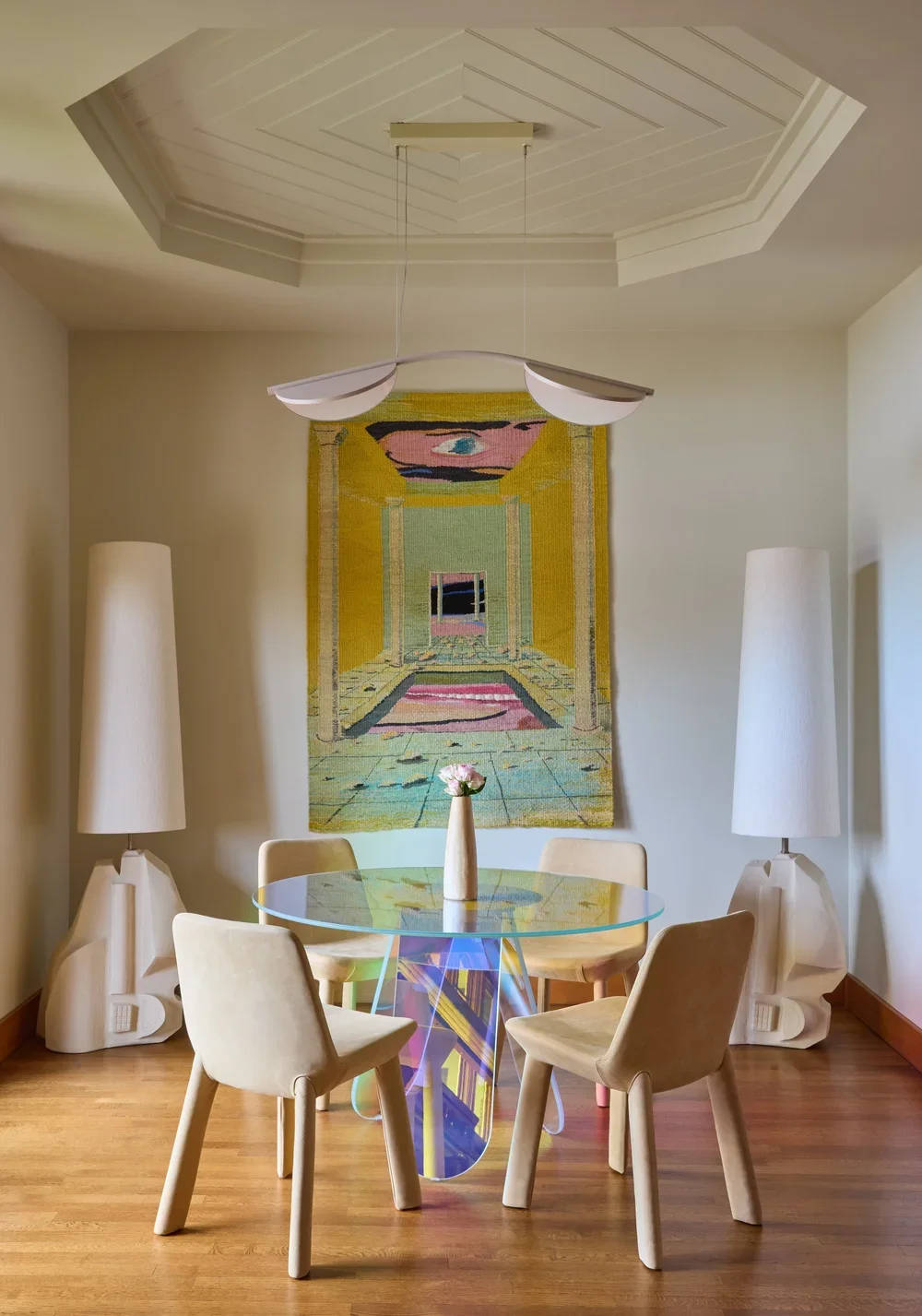 Modern dining space featuring a round glass table with an iridescent sculptural base, surrounded by minimalist beige chairs. The room is accented with contemporary floor lamps and a bold, colorful wall tapestry, creating a balance of artful design an