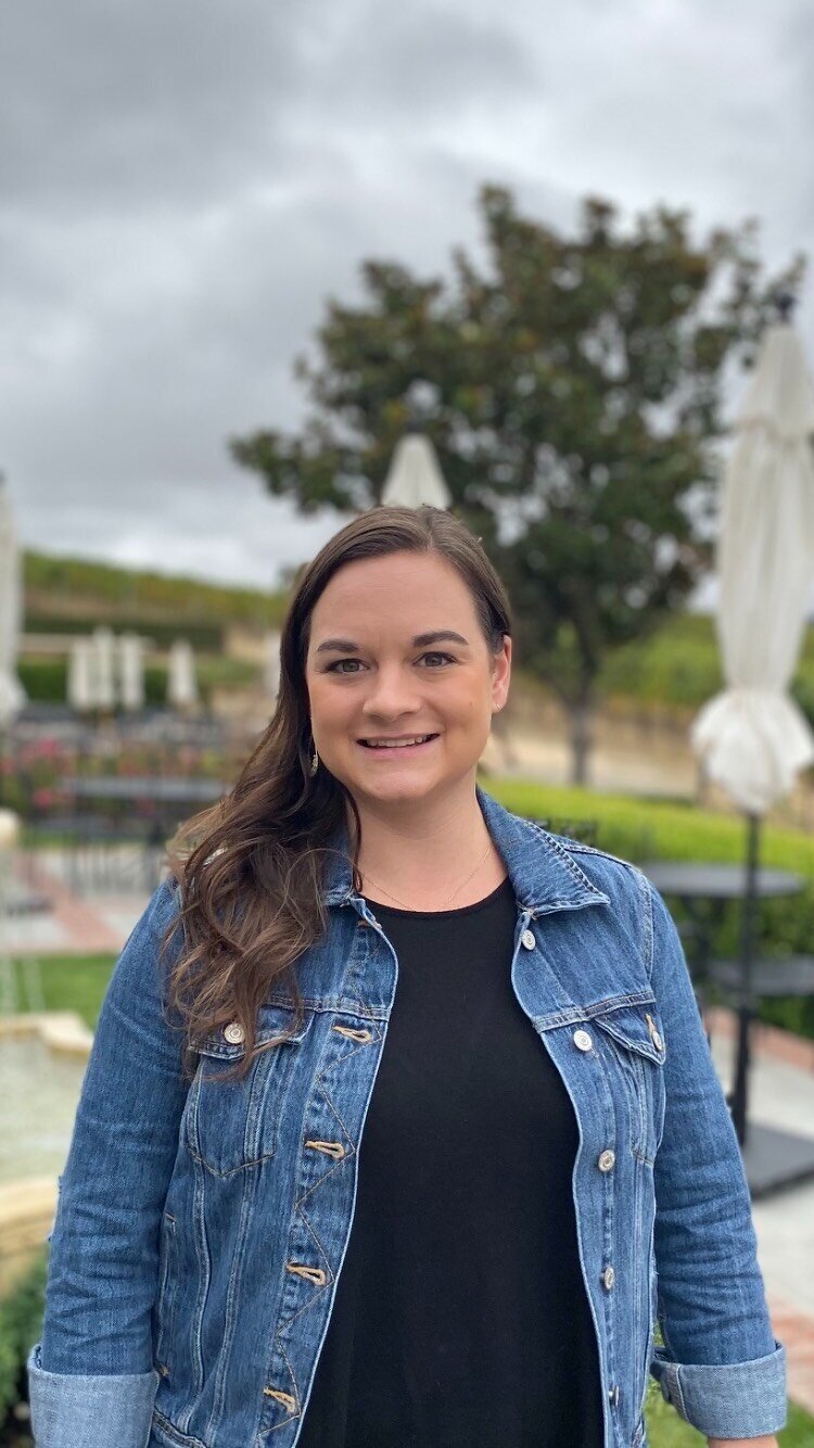 A smiling woman with long brown hair wearing a denim jacket and black shirt outdoors on a cloudy day. Therapy for Women in Bend, OR - Bend Thoughts & Therapy.