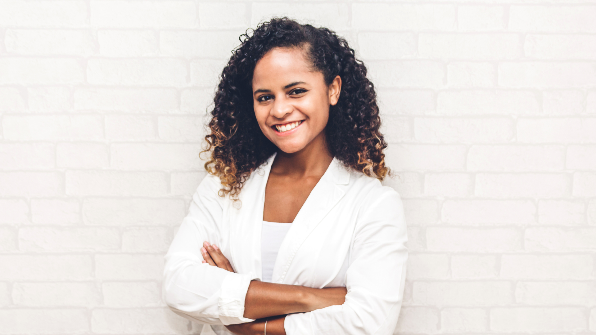 A young woman with curly hair smiling with arms crossed standing against a white brick wall. Bend Thoughts and Therapy accepts insurance.