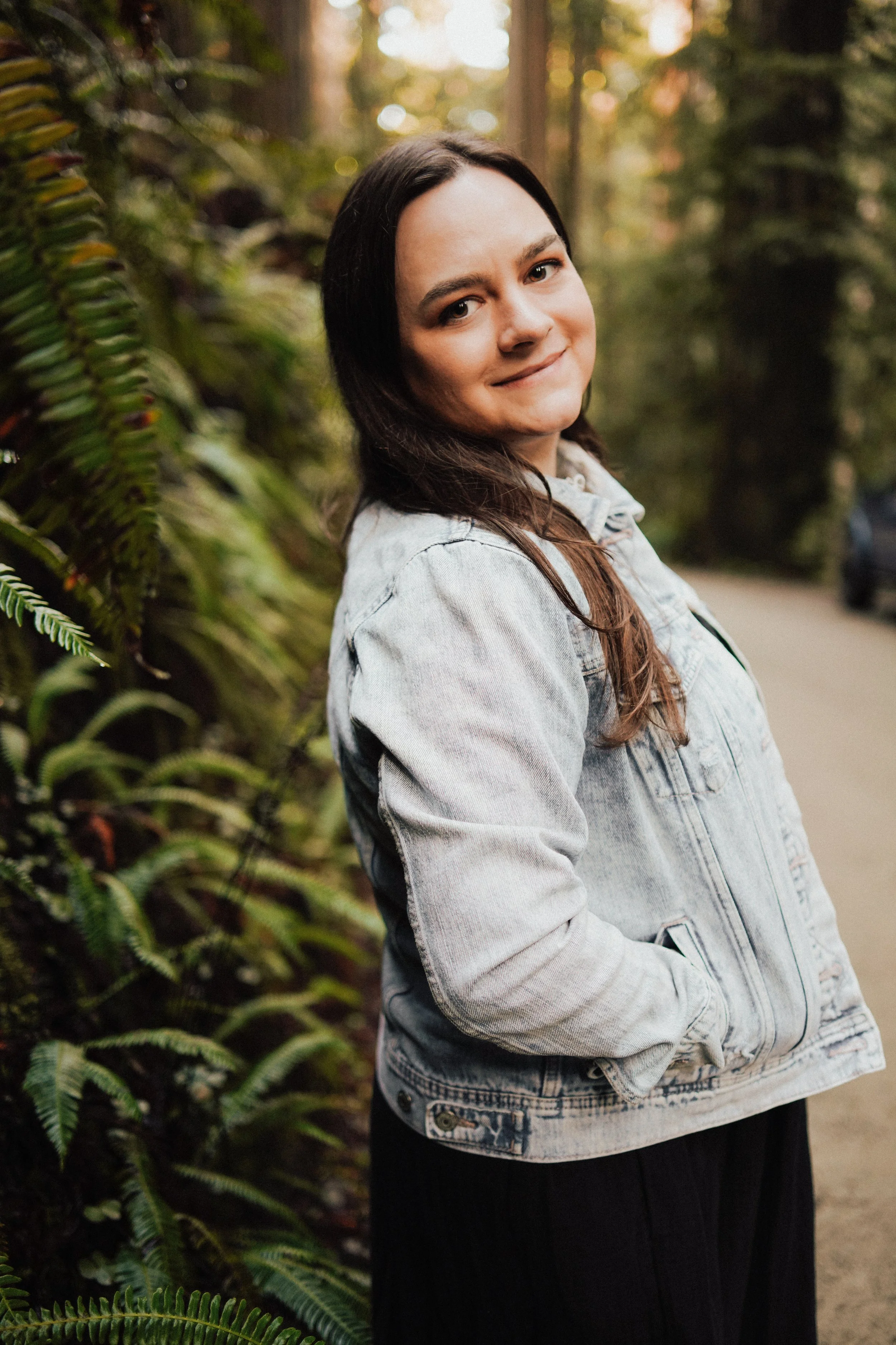 A young woman with dark hair smiling and standing outdoors in a forested area, wearing a light denim jacket and black pants. Therapy for Women in Bend, OR - Bend Thoughts & Therapy.