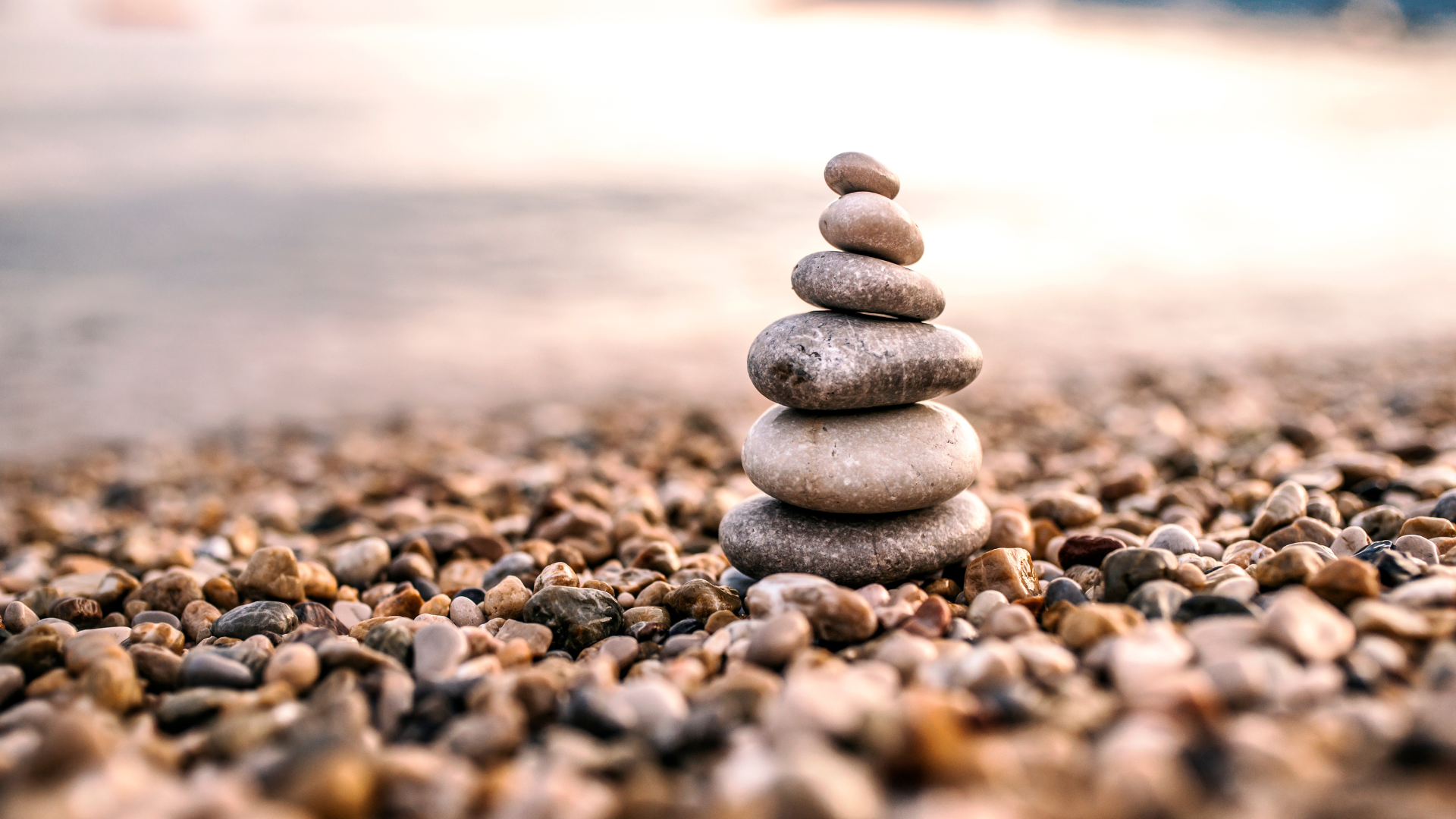 A stack of smooth, rounded stones arranged in a balanced tower on a pebble-covered beach with a blurred shoreline in the background.Bend Thoughts & Therapy in Bend, OR.