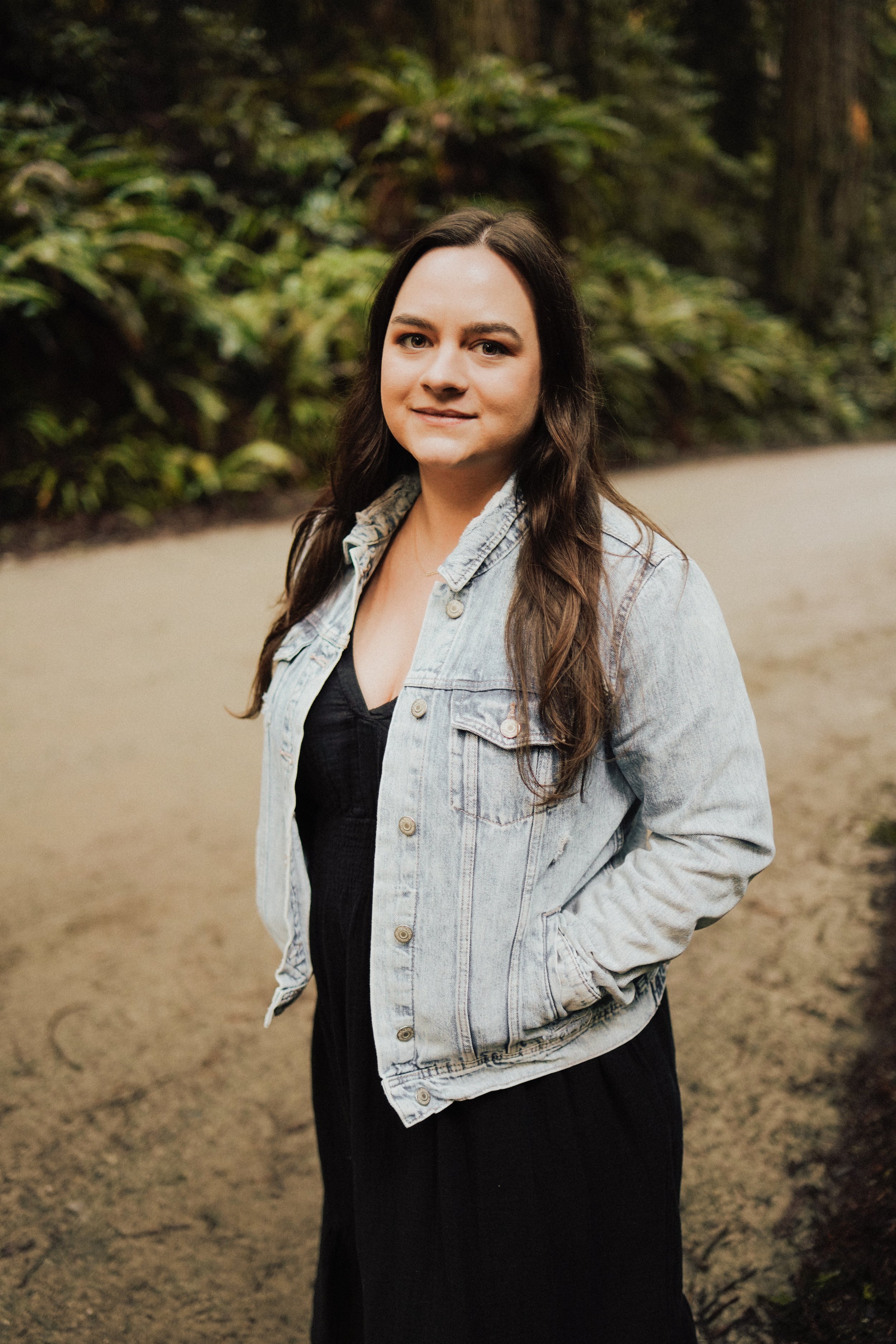 A young woman with long, wavy brown hair stands outdoors on a dirt path, wearing a denim jacket over a black dress, with greenery in the background. Therapy for Women in Bend, OR - Bend Thoughts & Therapy.