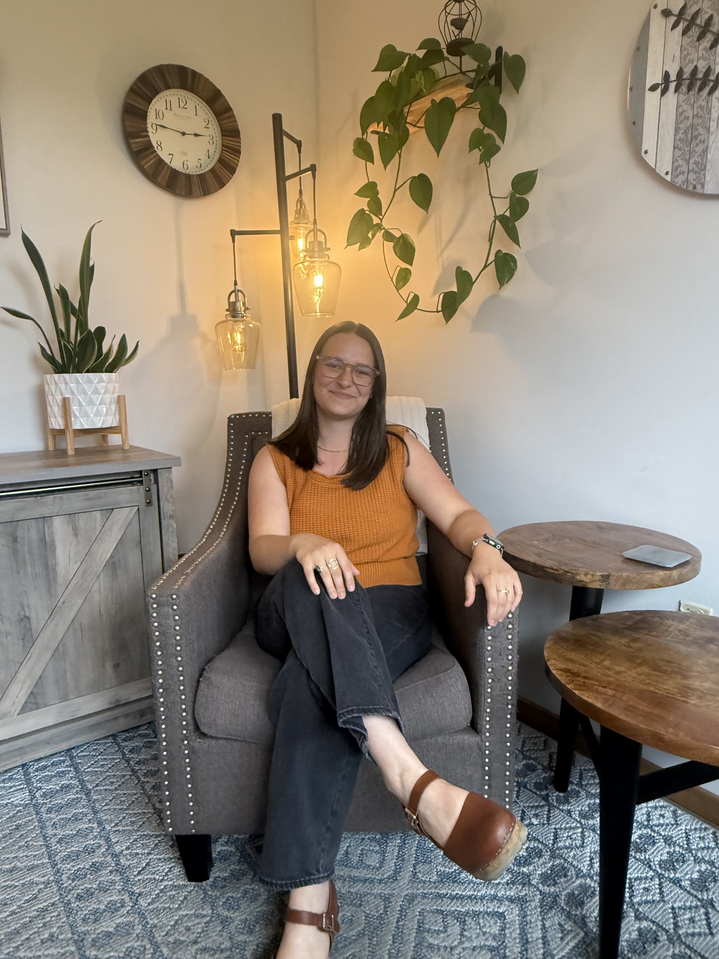 A woman sitting in a gray armchair in a cozy living room, wearing glasses, a sleeveless orange top, dark jeans with rolled-up cuffs, and brown shoes. She is smiling, with her arms resting on the armrests and her left leg crossed over her right.  Bend