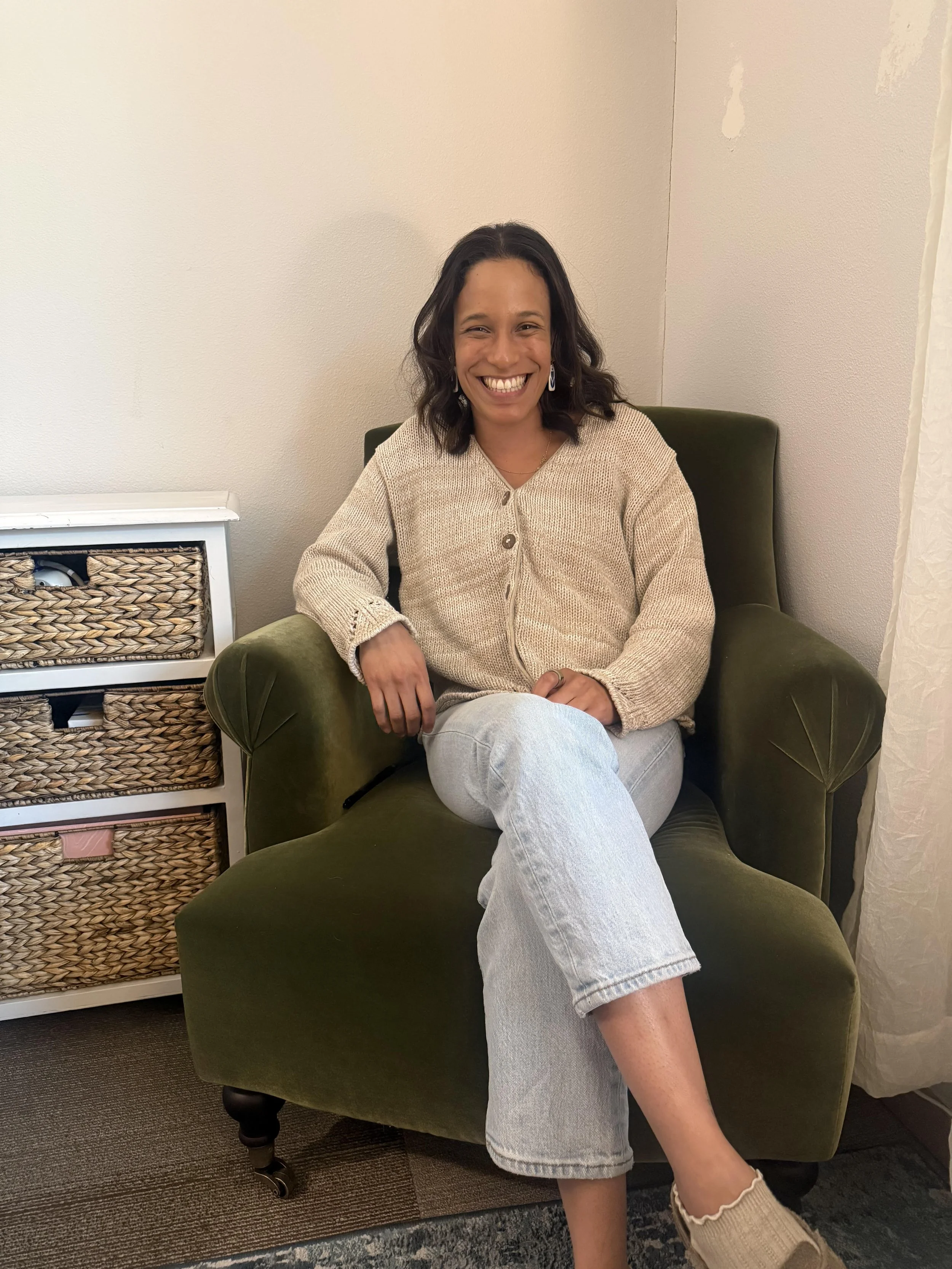 Khatren, a master’s level clinician with a degree in Clinical Mental Health Counseling from Bellevue University, sitting in a green armchair, smiling, wearing a beige knit cardigan, light jeans, and beige socks, next to a woven basket storage unit.