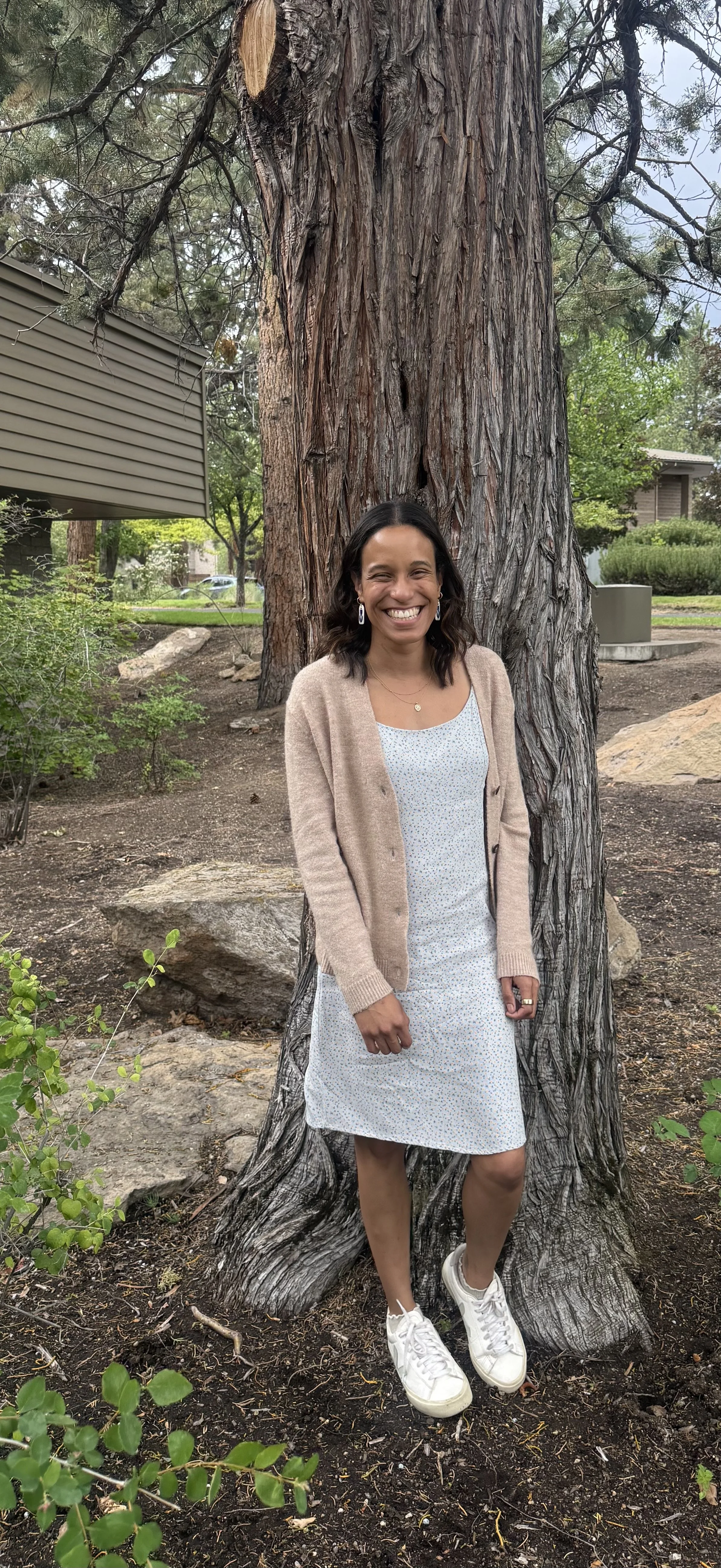 A woman standing in front of a tree in a park, smiling at the camera. She is wearing a light-colored dress, a beige cardigan, white sneakers, and earrings. The scene is outdoors with greenery and rocks in the background.  Bend, OR therapy