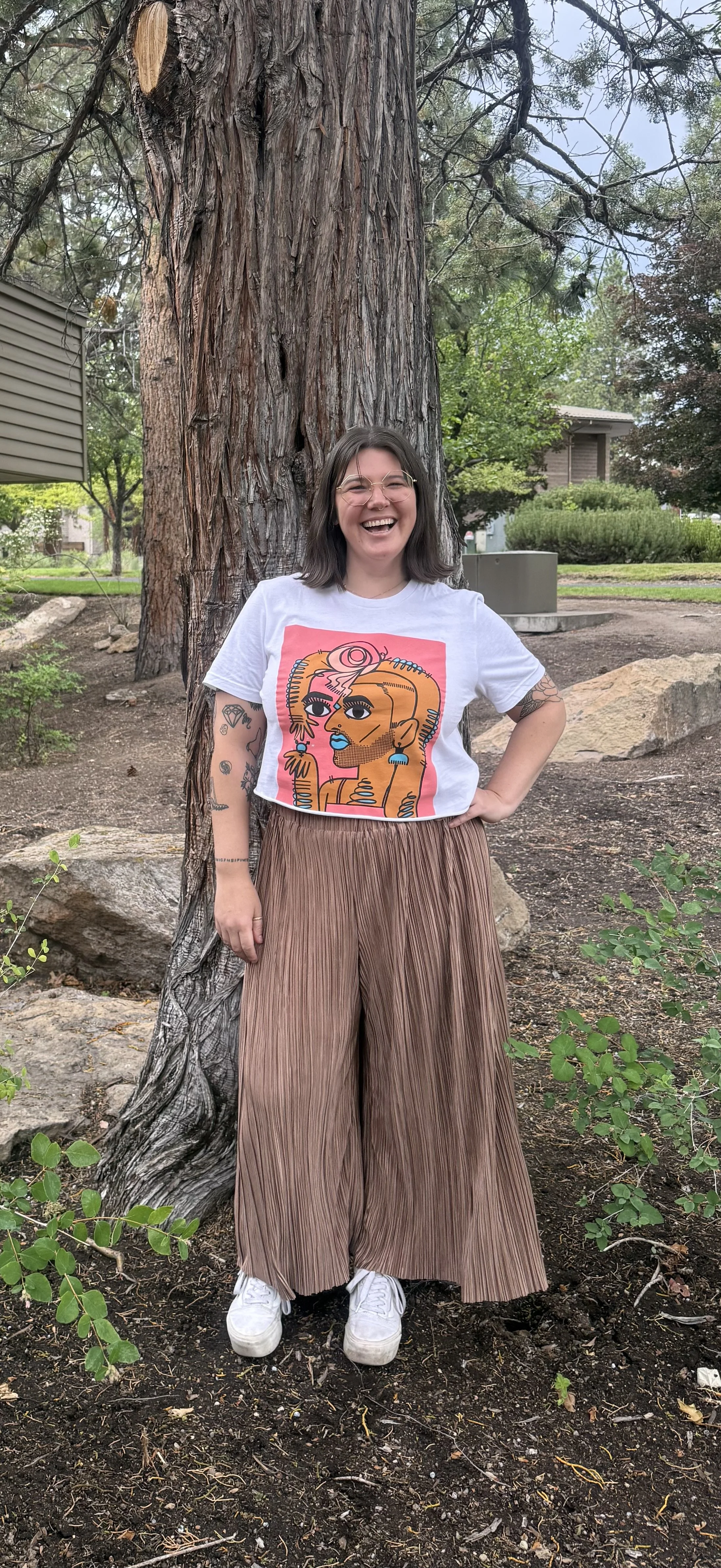 A woman with shoulder-length dark hair, glasses, and tattoos on her arms, standing outdoors next to a large tree, wearing a white graphic T-shirt, wide brown pleated pants, and white sneakers, smiling at the camera. Bend, OR therapy
