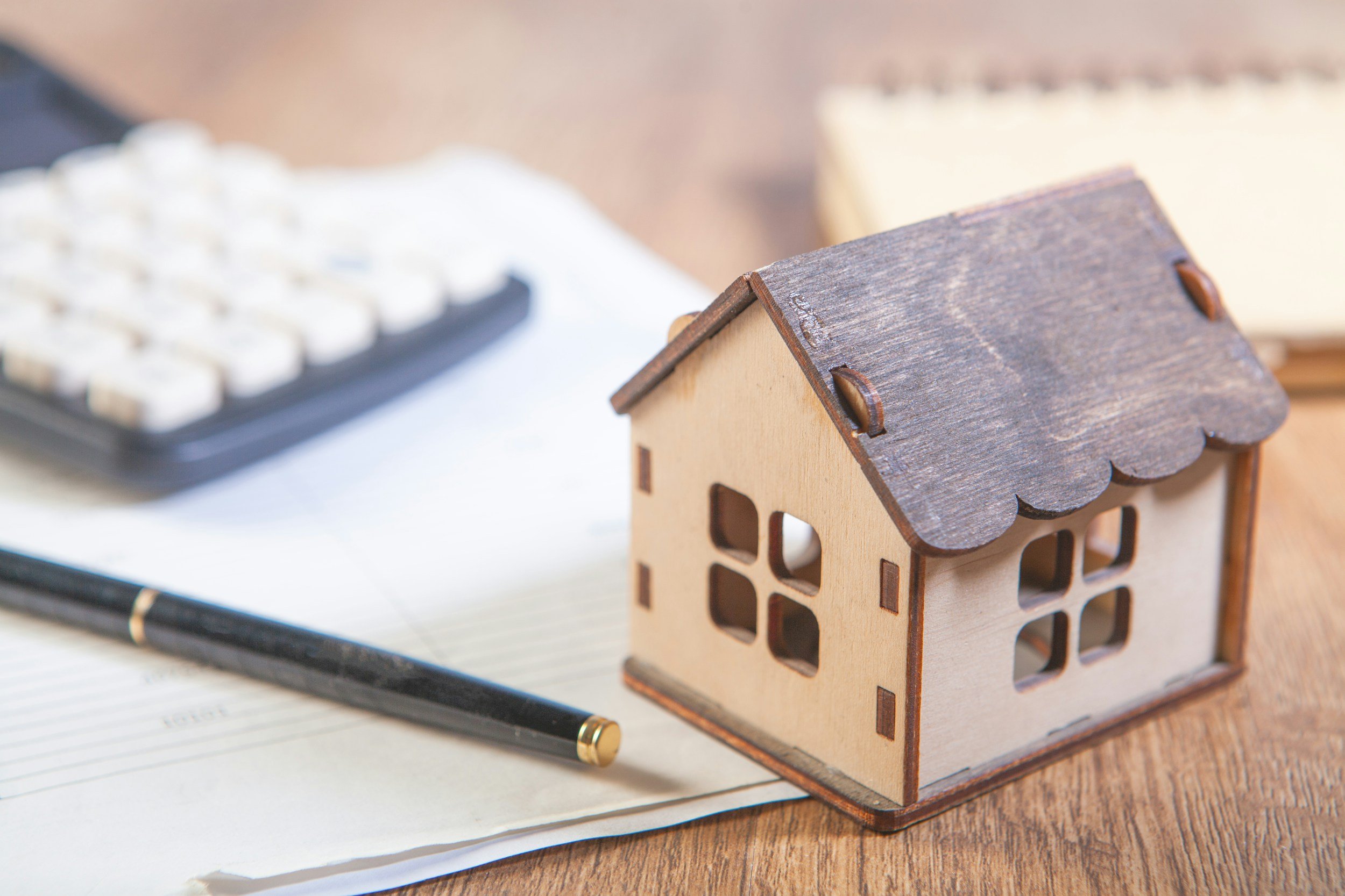 Several items on a table, including a small wooden model of a house, a pen, and lined paper. There is a keyboard and a notepad out of focus in the background.