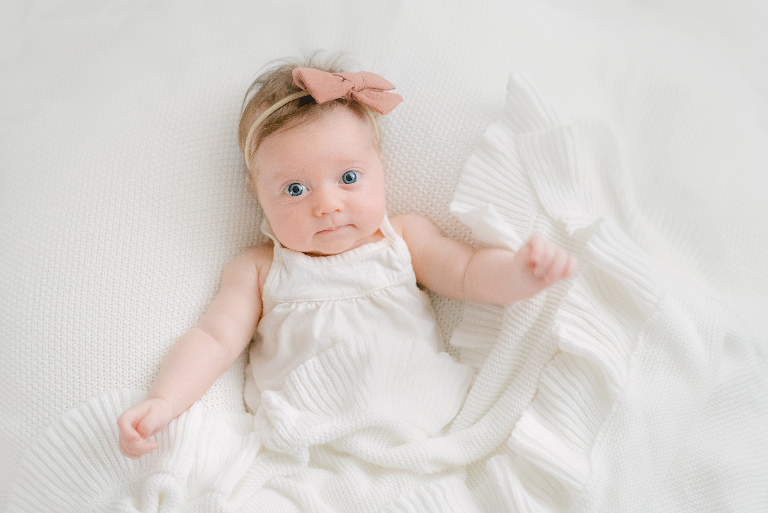 Baby girl with blue eyes and pink bow lying on white knit blanket, highly recommended baby photographer in Orange County