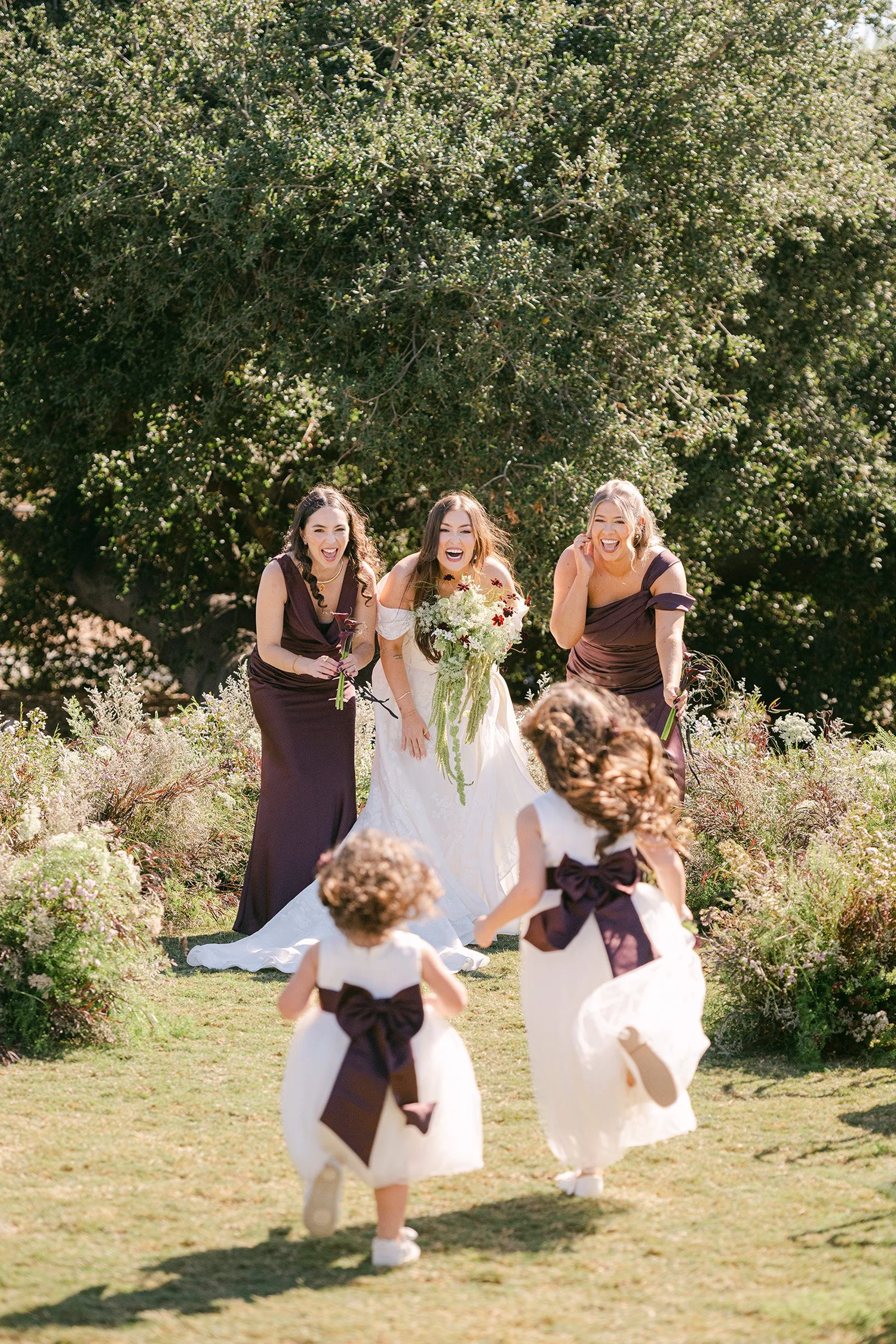 Orange County wedding photographer captures joyful bride with bridesmaids in plum gowns as flower girls run through the garden of The Swanner House Wedding Venue in San Juan Capistrano