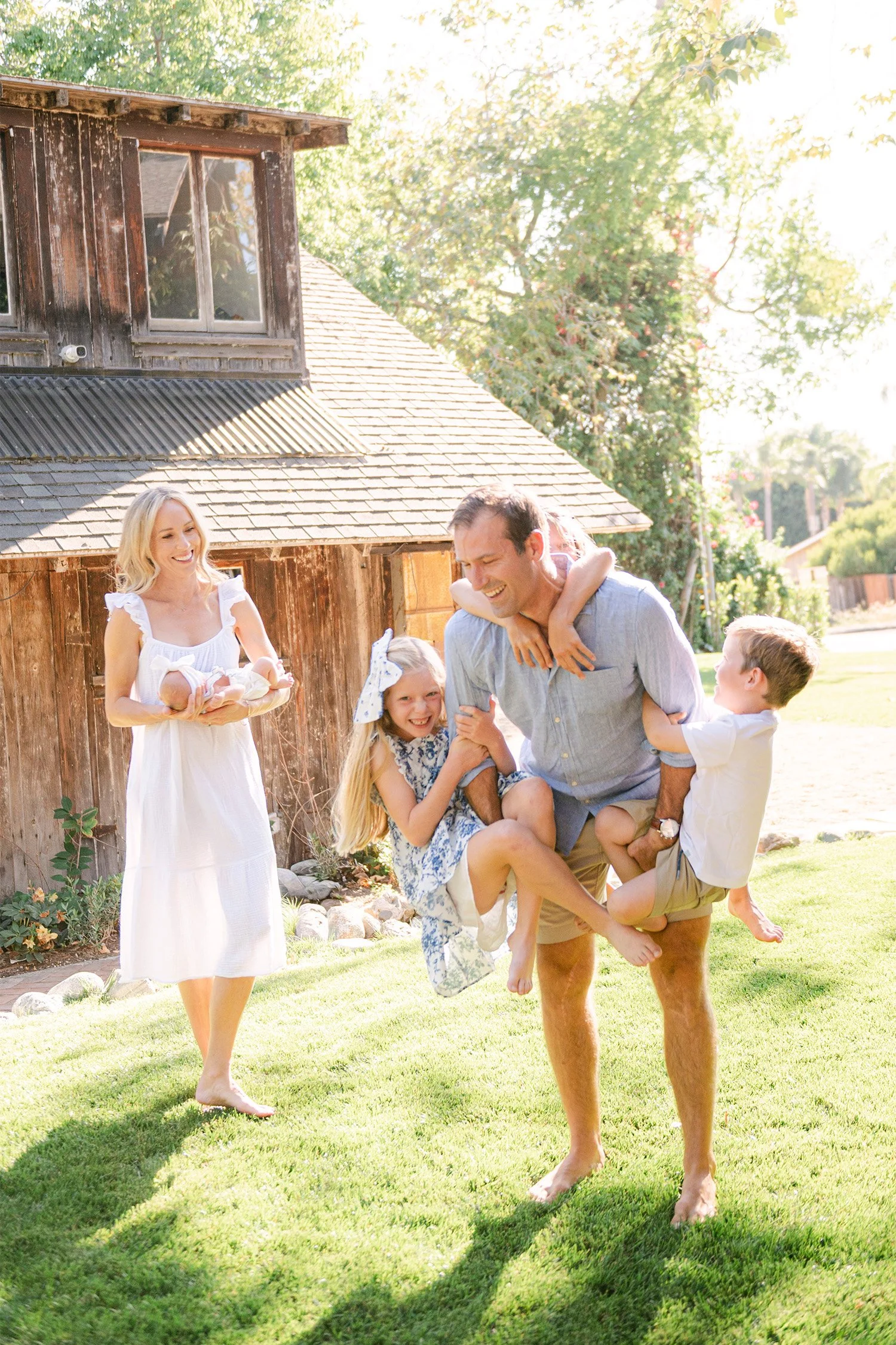 Father carrying laughing kids while mother holds newborn by a barn, Orange County family photographer