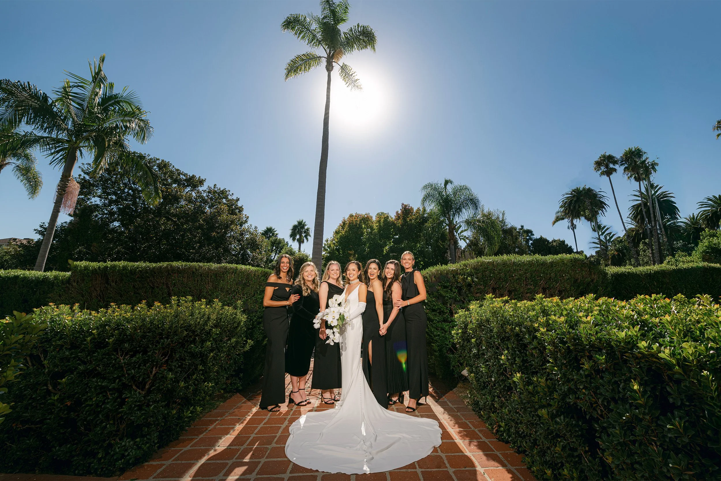 Award-winning Orange County wedding photographer captures bride with bridesmaids in black dresses posing under palm trees on garden pathway in Santa Barbara
