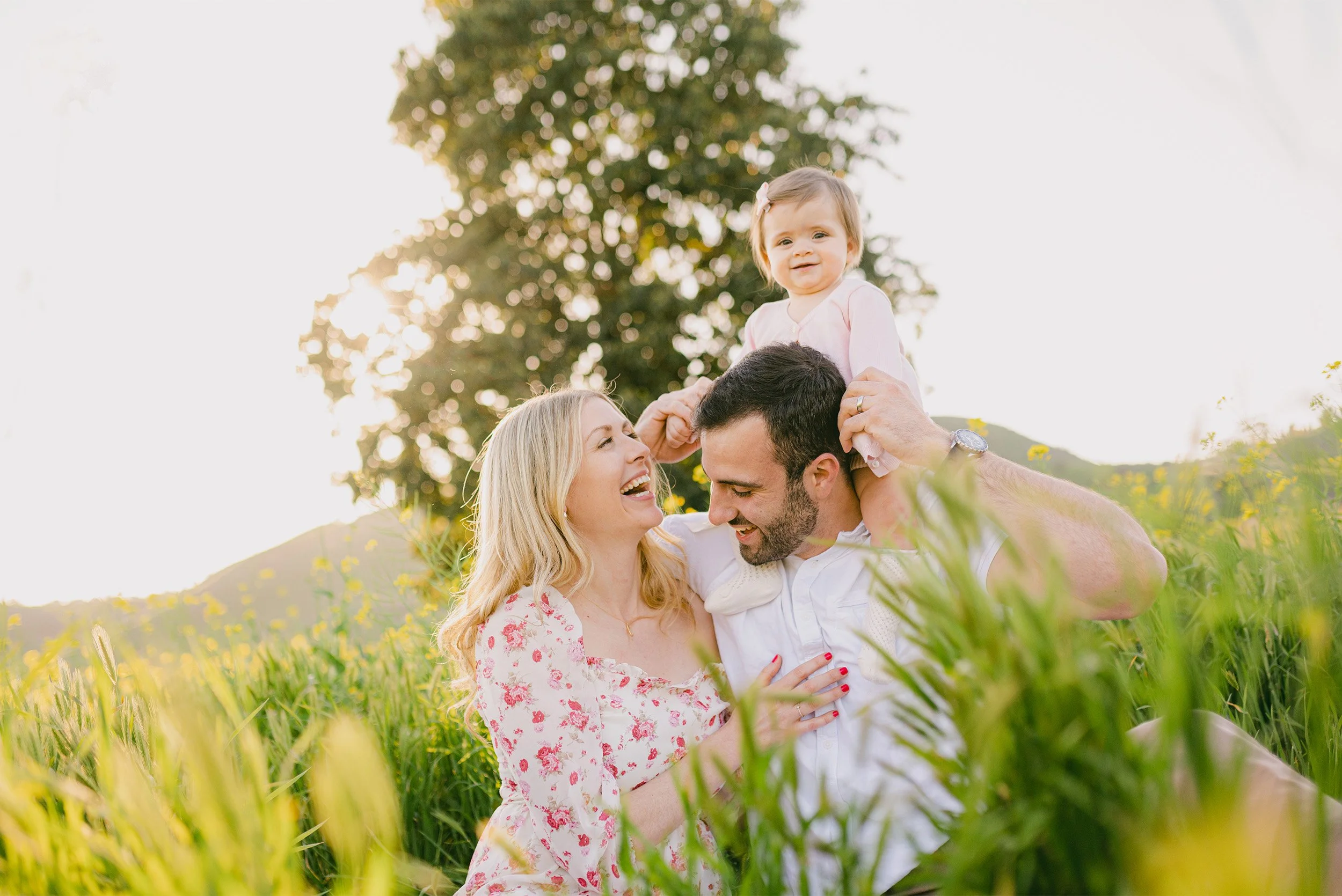 Orange County family photographer captures parents laughing with daughter on shoulders in green field
