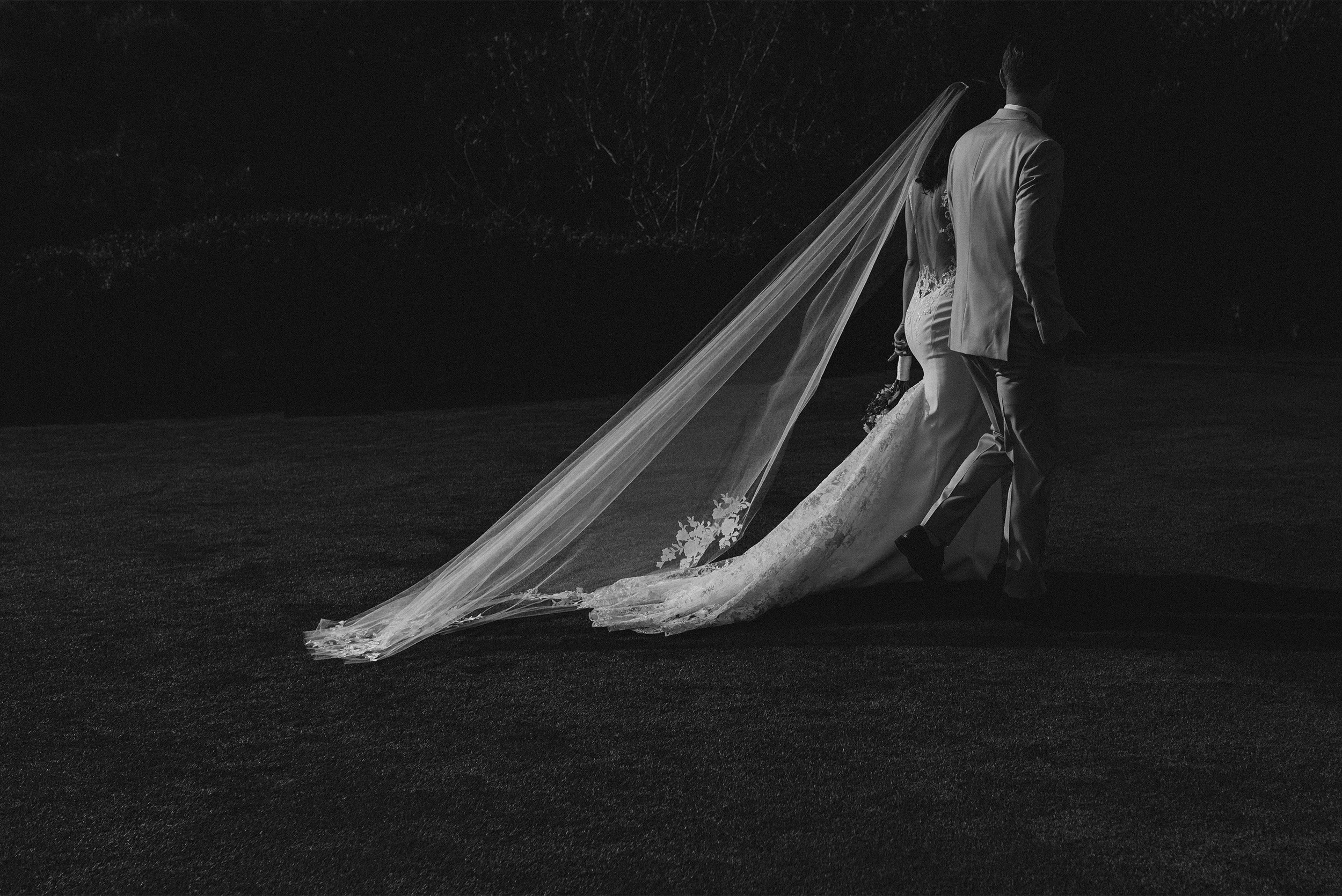 Award-winning wedding photographer in Santa Barbara captures newlyweds walking hand in hand with cathedral veil trailing, black and white