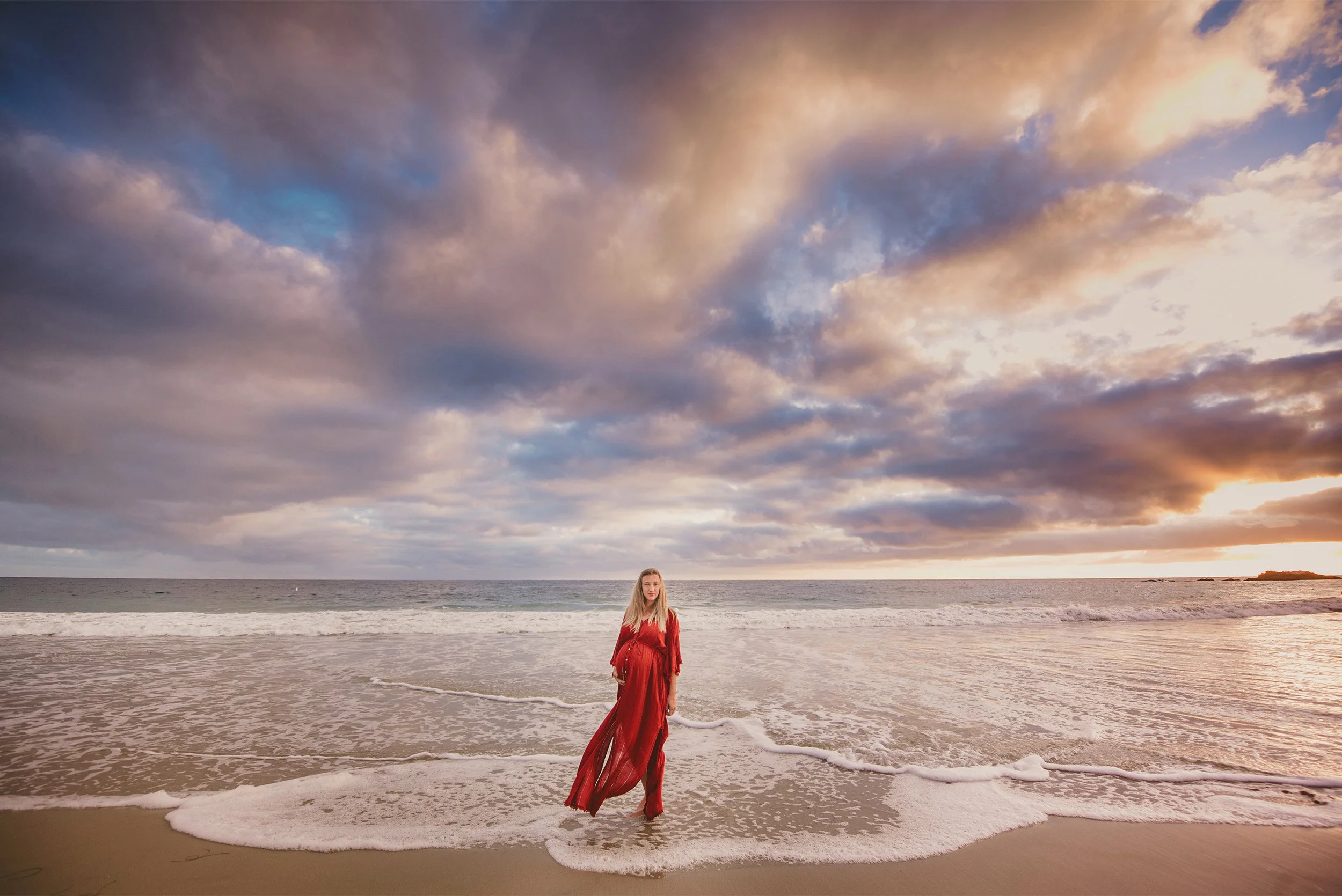 Orange County maternity photographer photographs pregnant woman in red dress standing in ocean waves at sunset