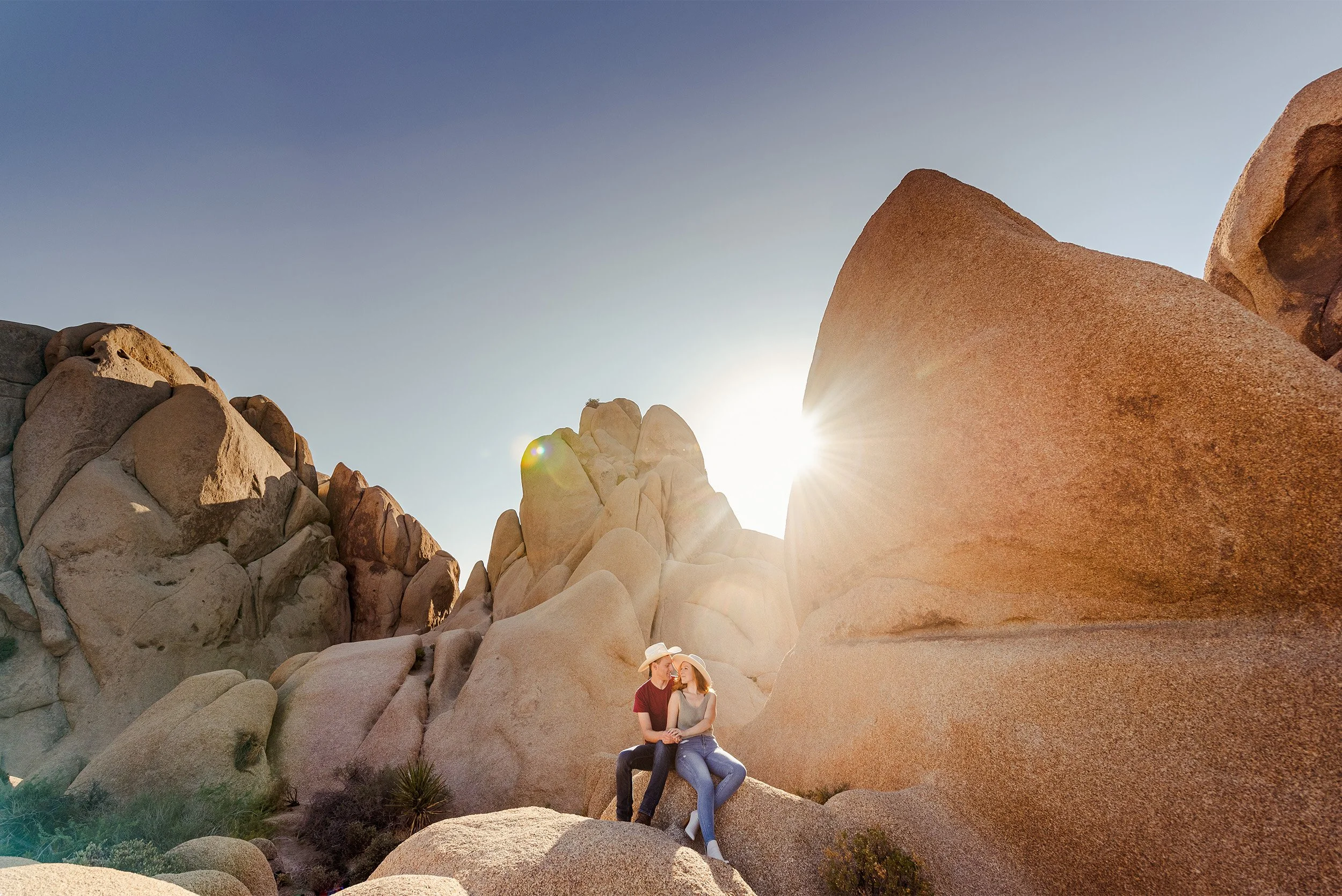 Couple in cowboy hats sitting together among the boulders of Joshua Tree National Park at sunset for an engagement announcement