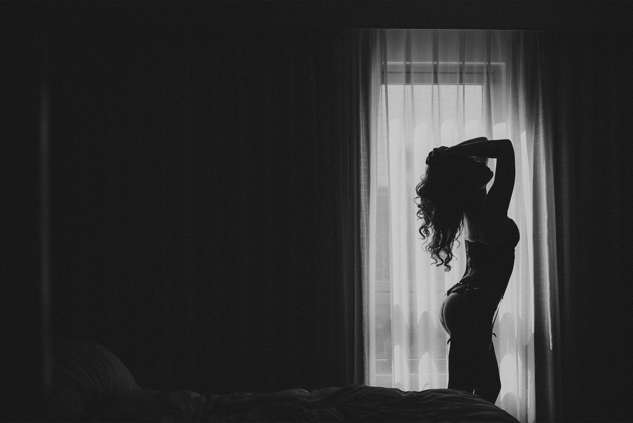 Orange County boudoir photographer captures silhouette portrait of woman backlit by sheer curtains in bedroom window light