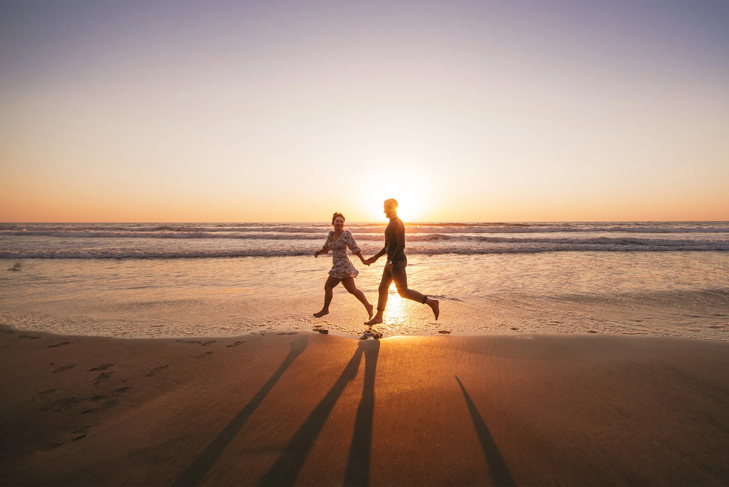 Couple running hand in hand along the shoreline at sunset, beach engagement photo session Southern California