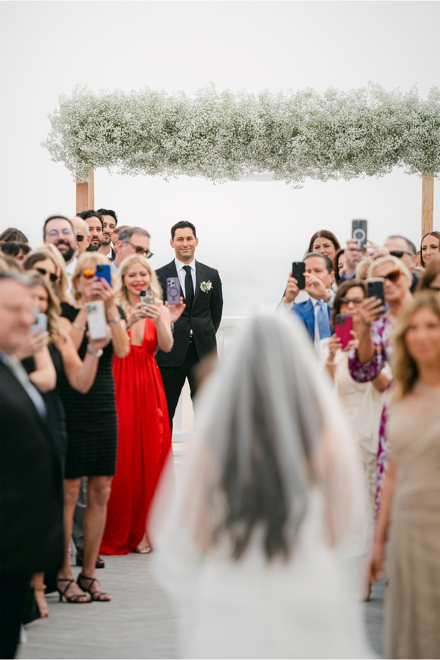 Groom smiling at bride walking down aisle as guests take photos, beachside wedding ceremony