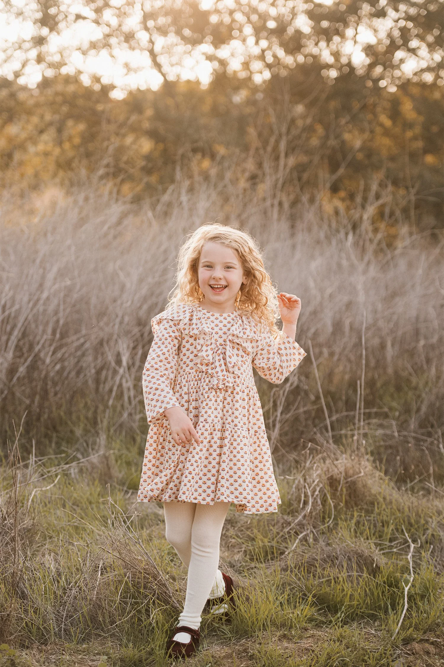 Curly-haired girl laughing in a floral dress at golden hour, Highly recommended family photographer photographs in Orange County