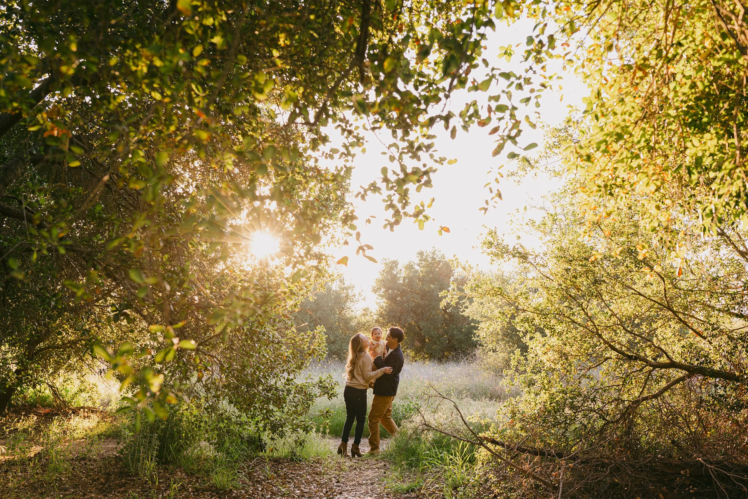 Orange County family photographer captures parents holding daughter in sun-dappled forest clearing