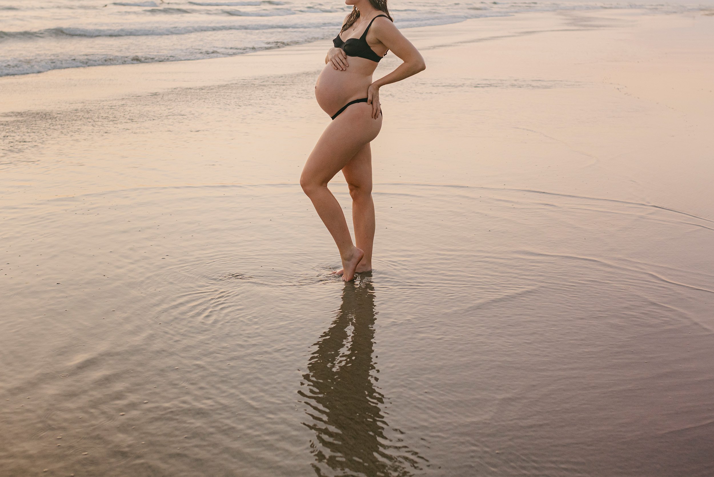 Close-up of pregnant belly in black bikini standing in shallow water, Orange County maternity photographer