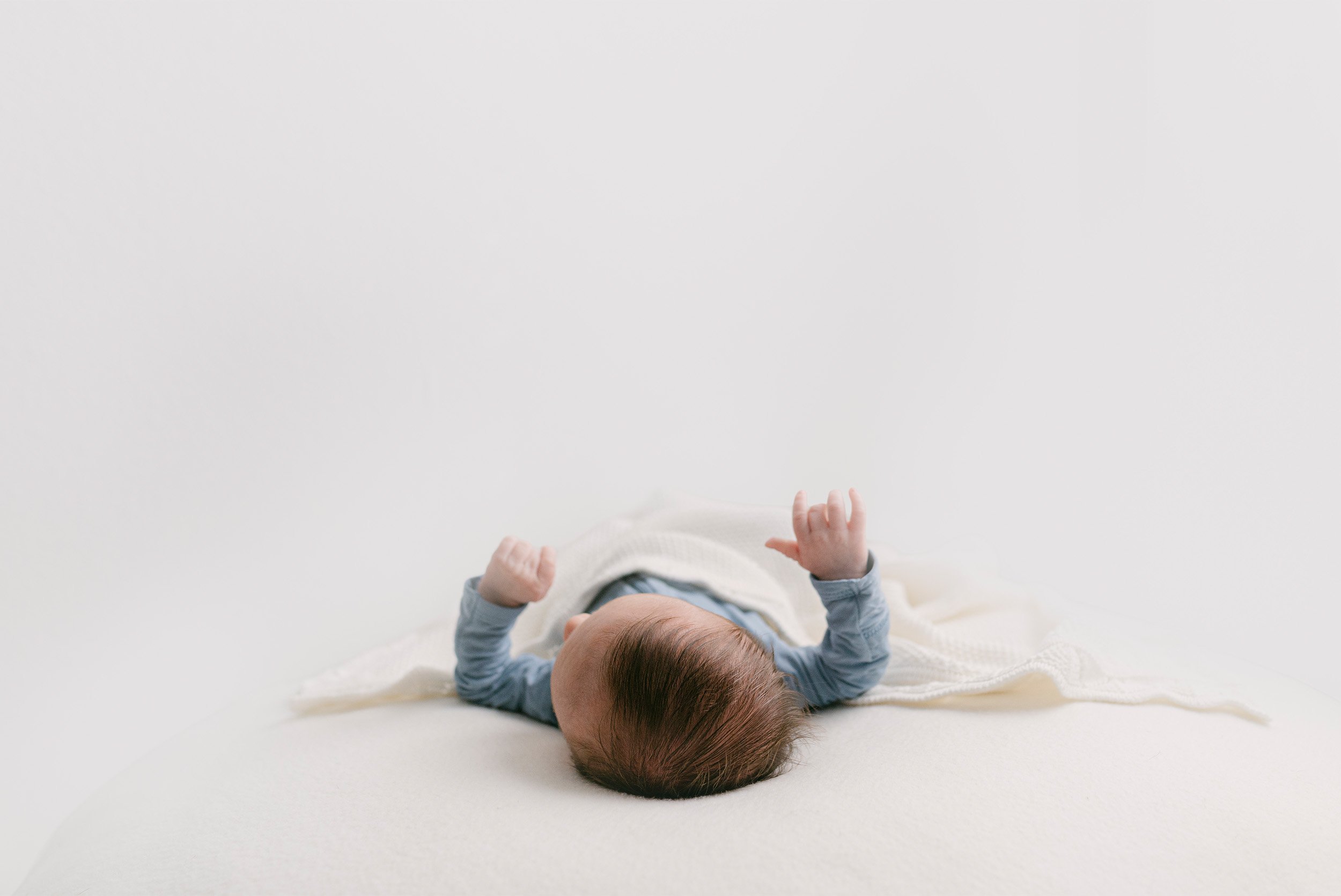 Orange County baby photographer captures newborn boy reaching up under soft white blanket during studio session, in home newborn photos