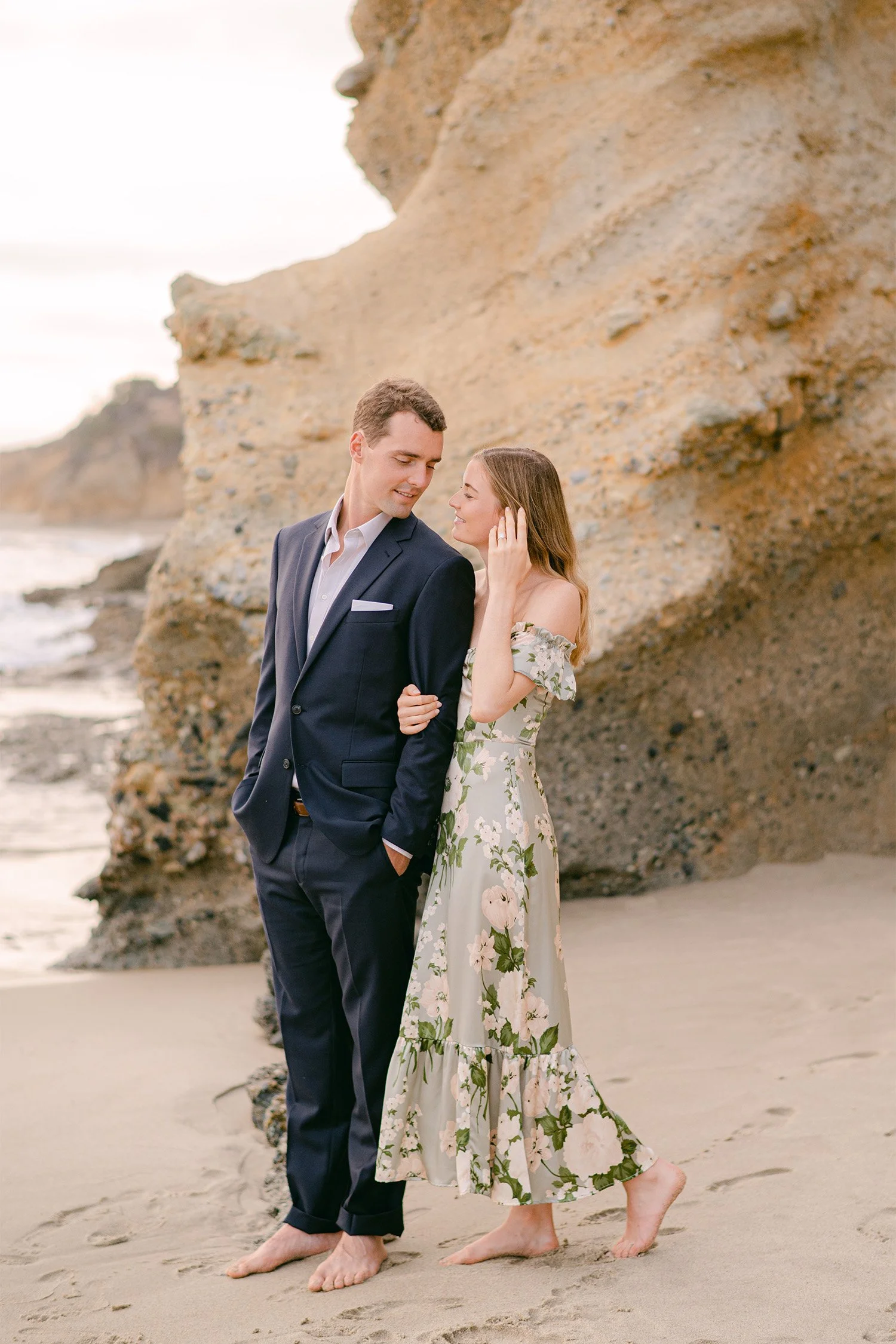 Highly recommended engagement photographer in Orange County captures couple standing close beside sandstone rock formation on the beach