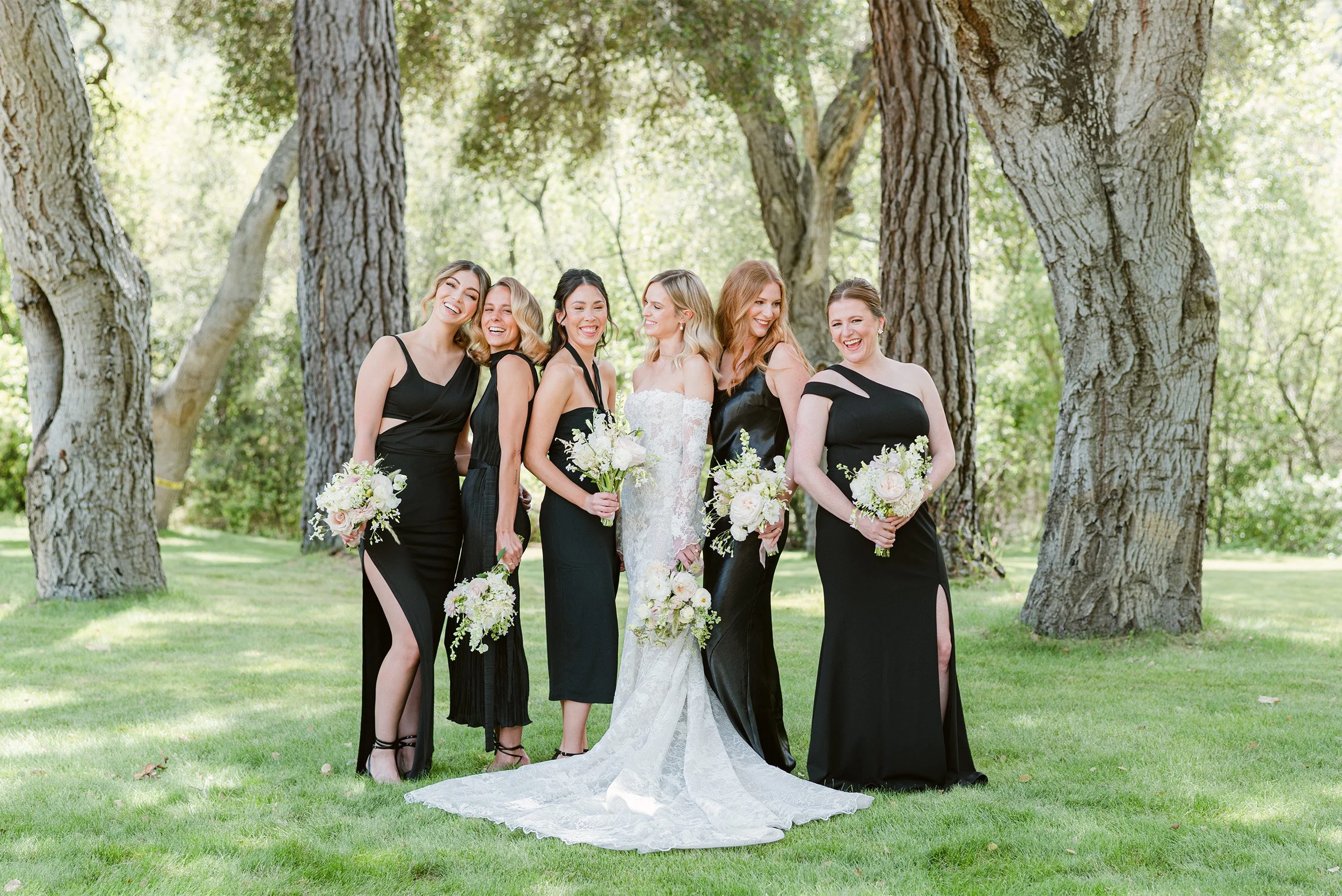 Orange County wedding photographer captures bride laughing with bridesmaids in elegant black dresses beneath towering oak trees at the Gardener Ranch in Carmel Valley