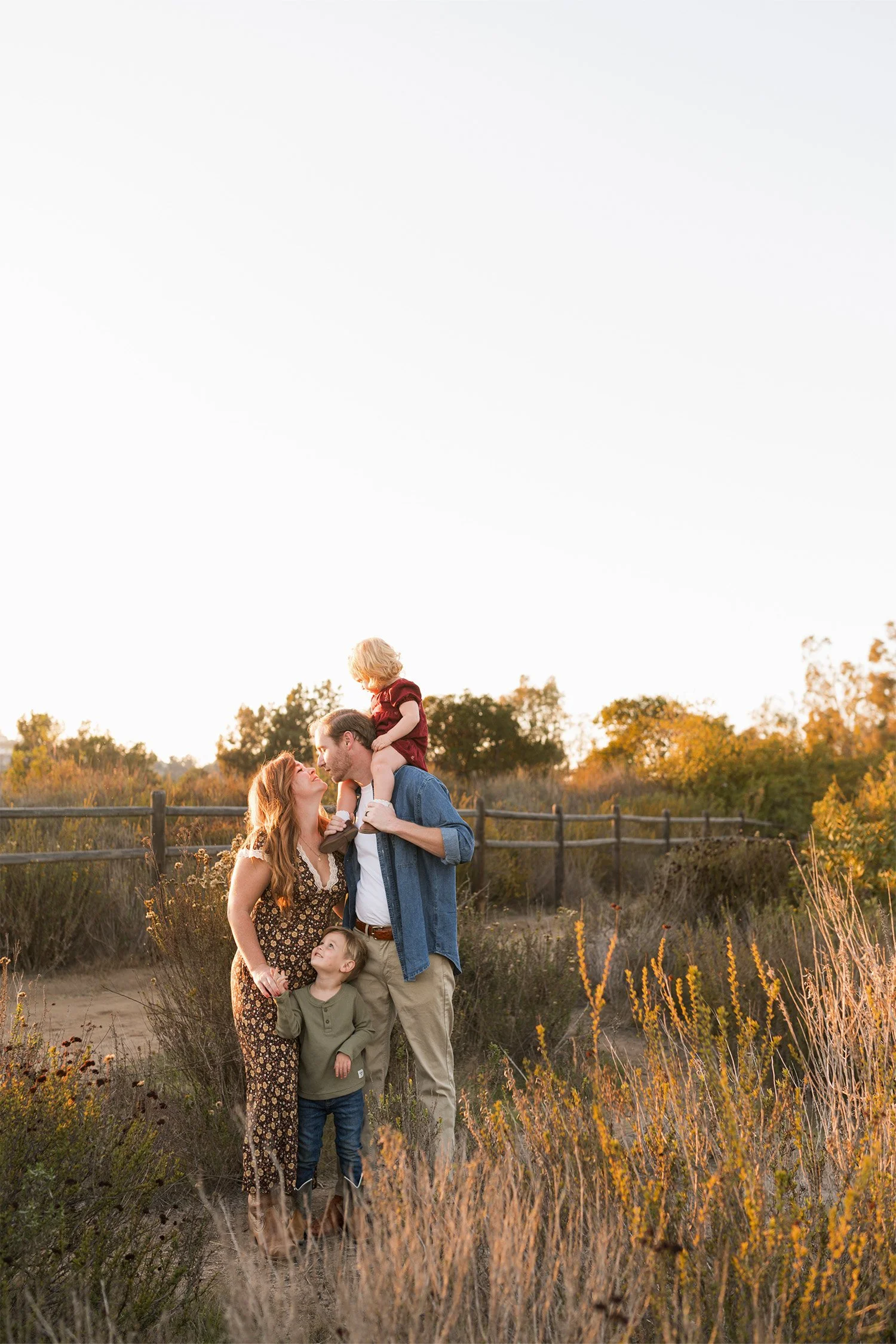 Parents and two children sharing a moment in a golden wildflower field with a wooden fence in the background, Orange County family photographer