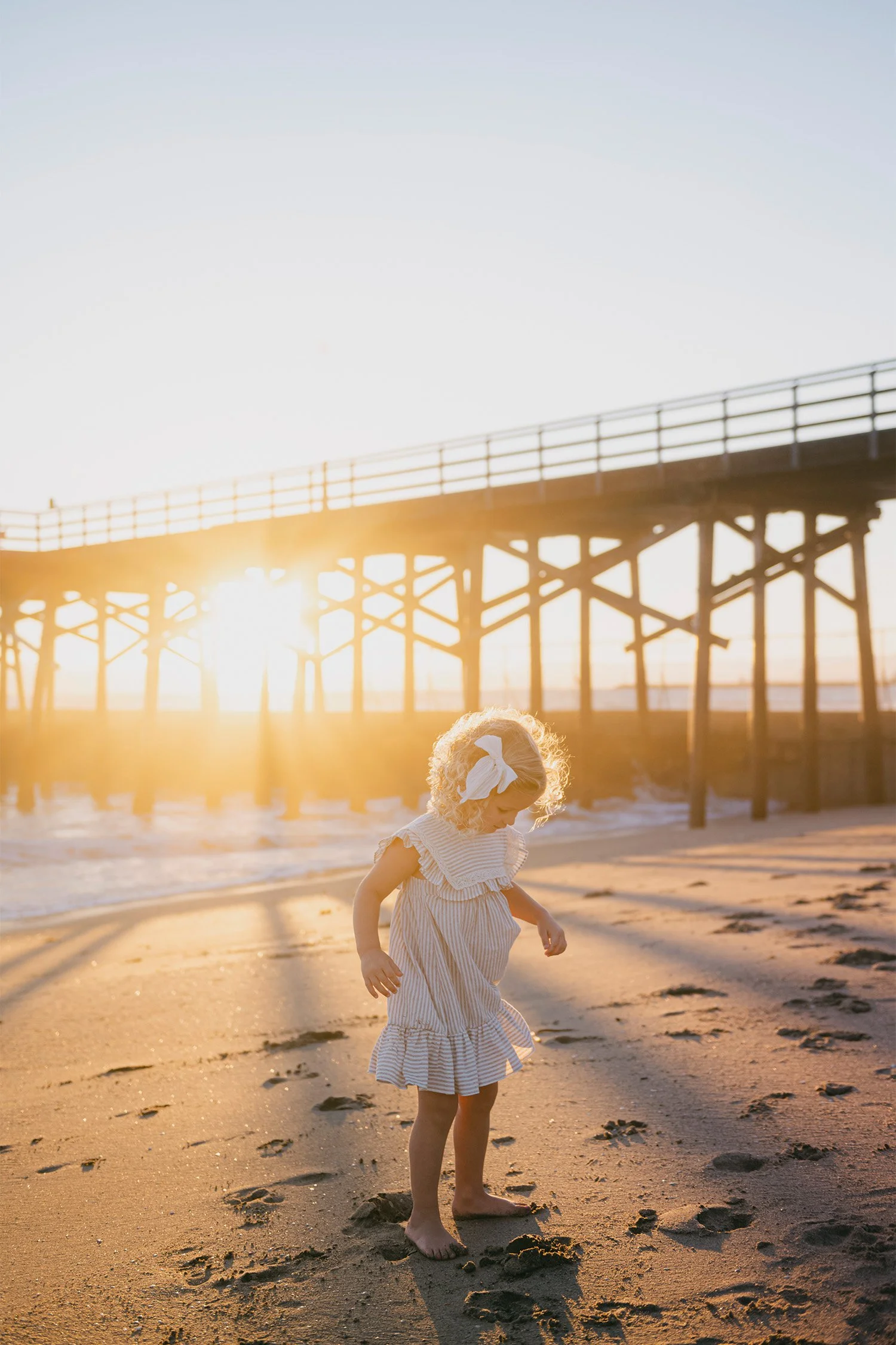 Family photography session of a curly-haired toddler girl playing on the beach near the Newport Pier at golden hour