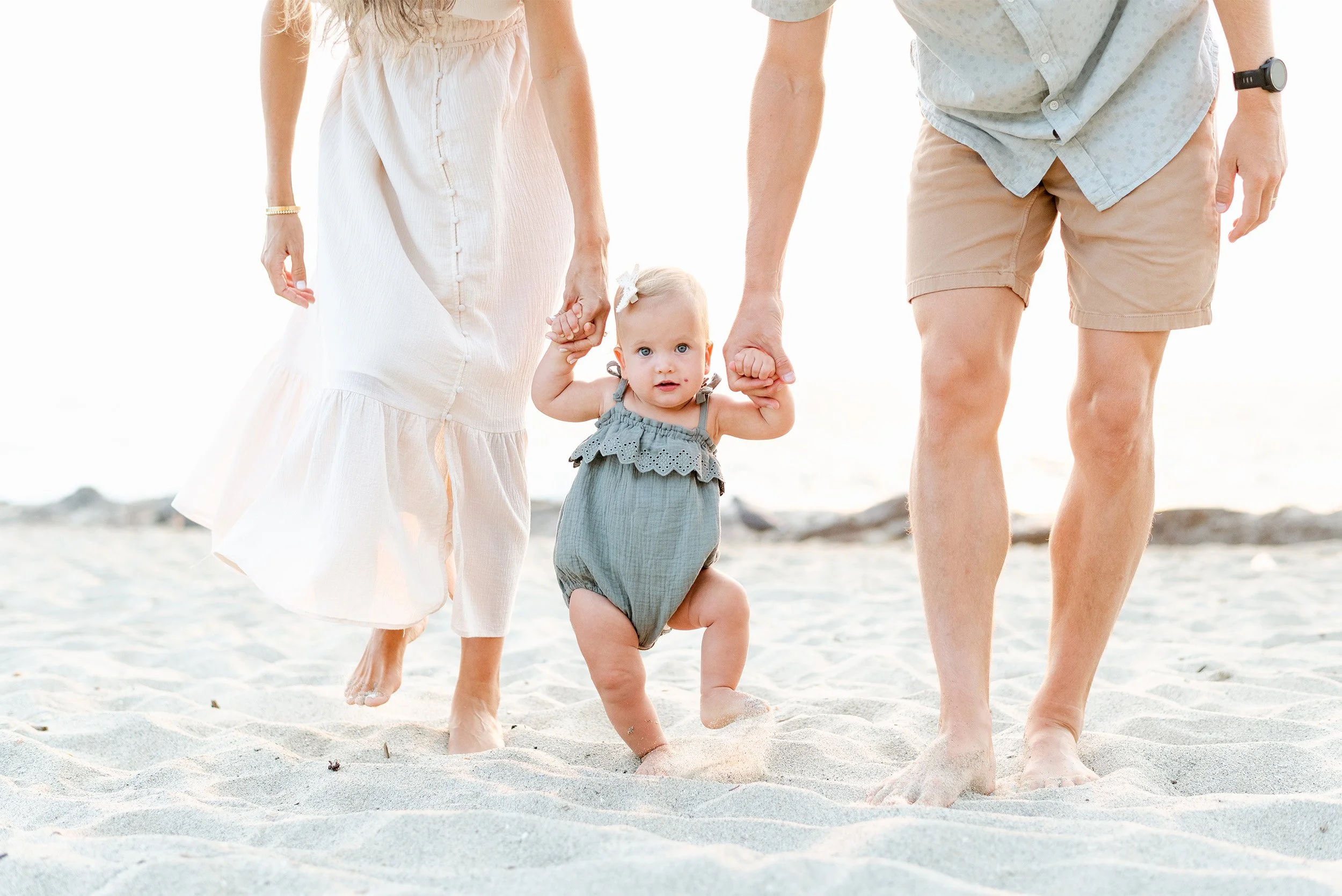 Award-winning family photographer in Orange County photographs toddler taking steps on the beach while holding her parents' hands