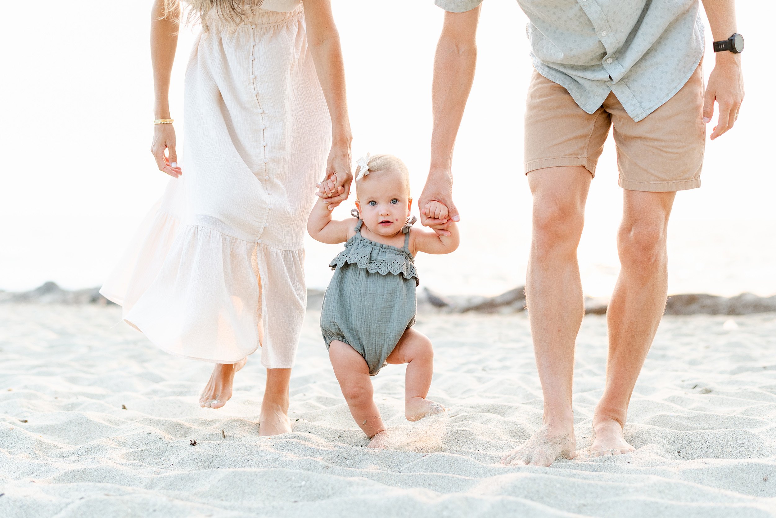 Award-winning family photographer in Orange County photographs toddler taking steps in Laguna Beach while holding her parents' hands