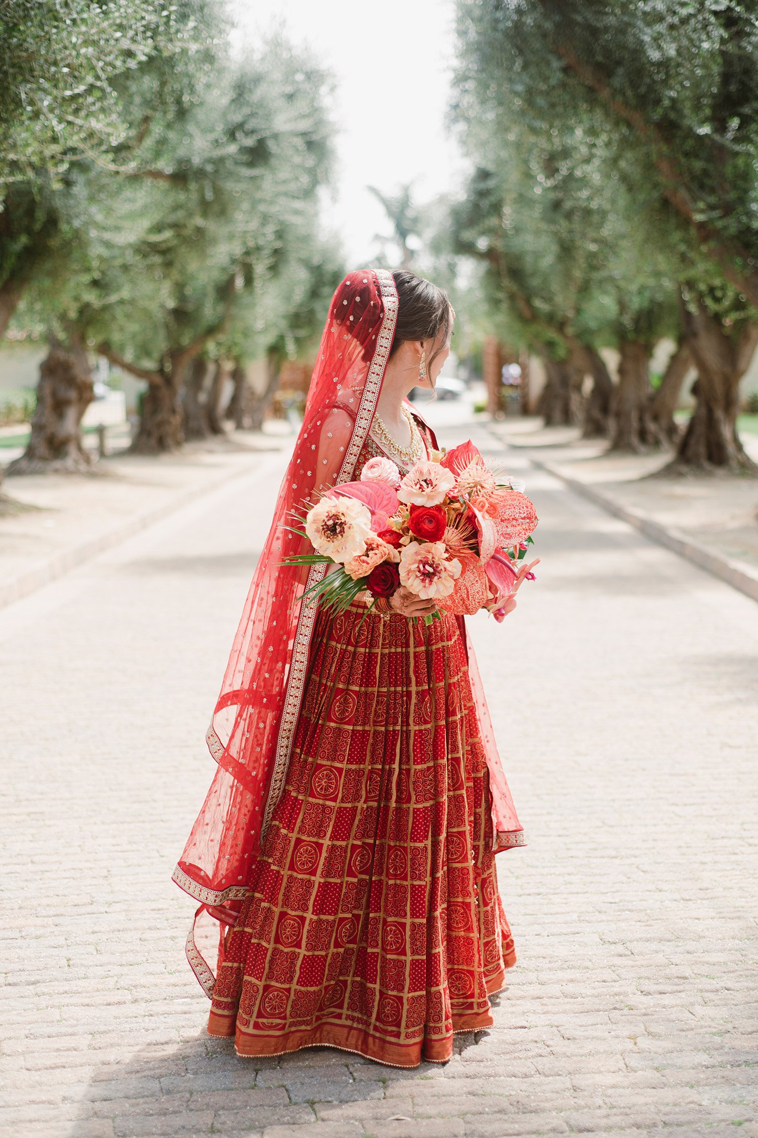 Highly recommended wedding photographer in Orange County captures bride in vibrant red lehenga walking beneath olive tree canopy
