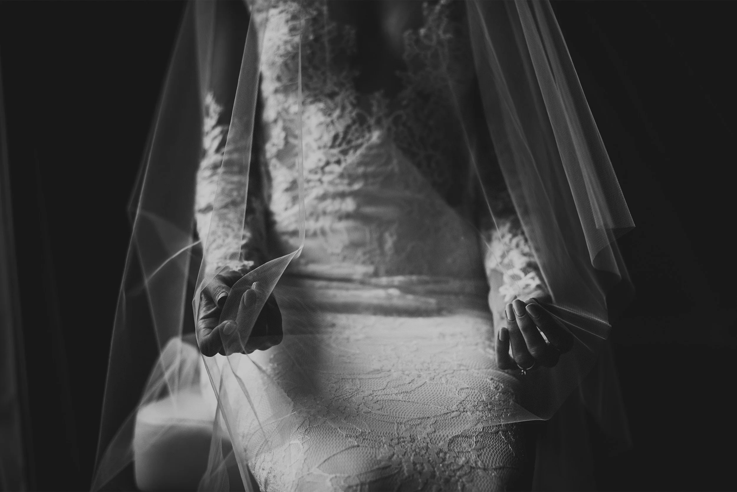 Bride's hands holding open flowing veil over lace wedding dress, black and white bridal detail photo