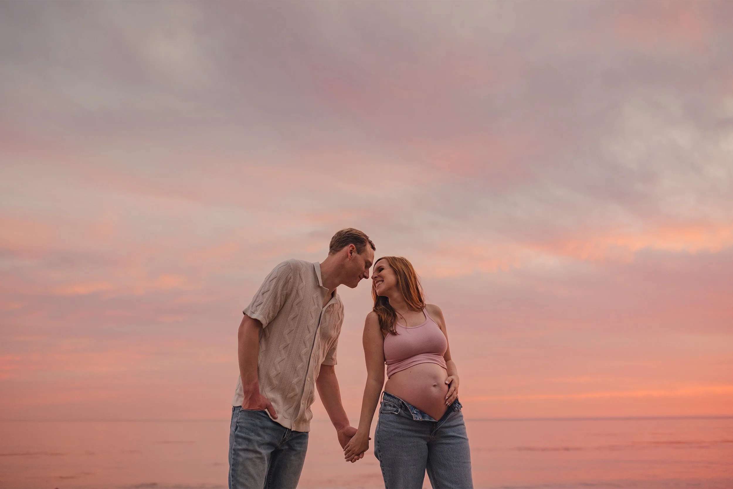 Couple touching foreheads on beach under pink sunset sky, award-winning maternity photographer in Orange County