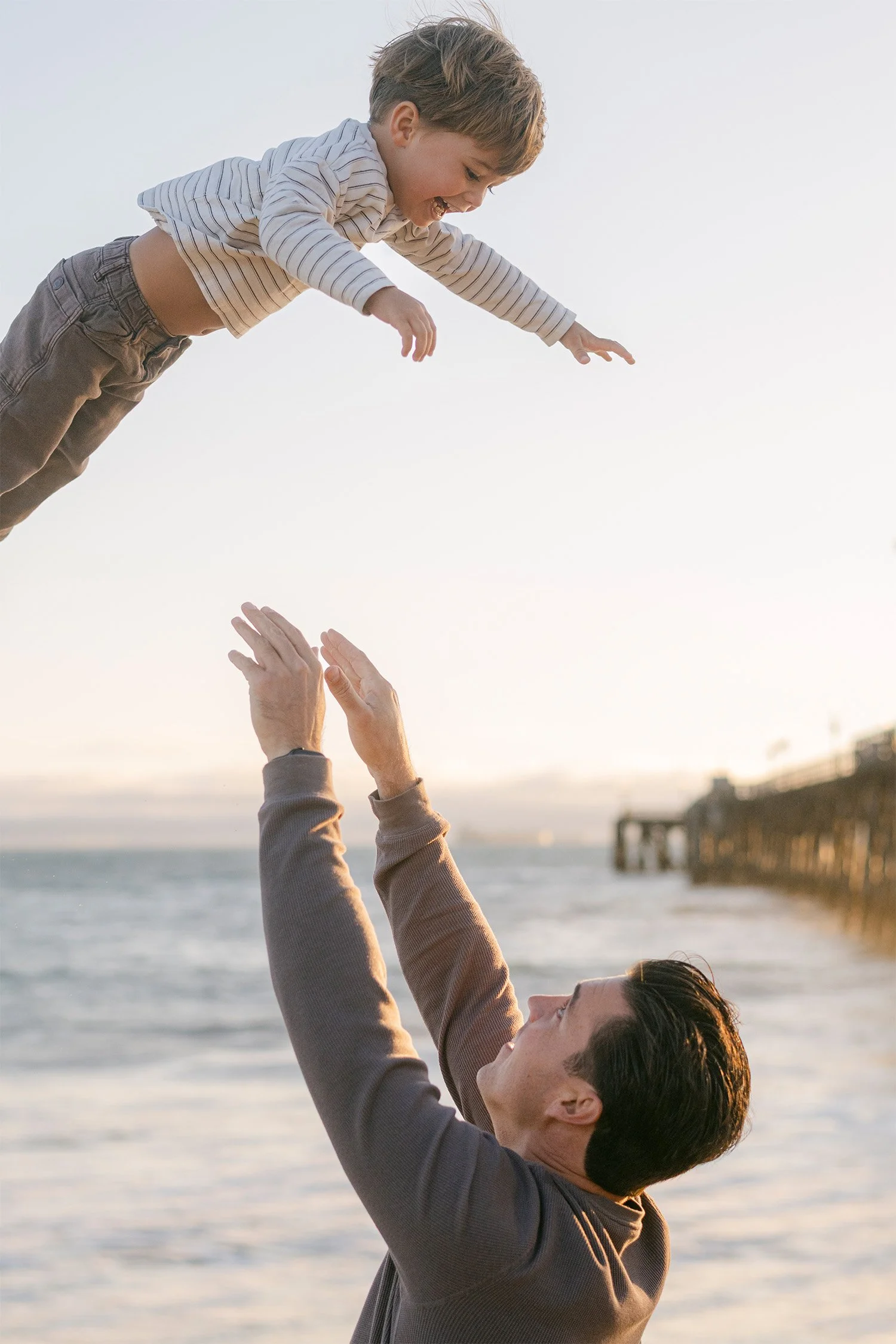 Father joyfully tossing his laughing son in the air at the beach, Orange County family photographer