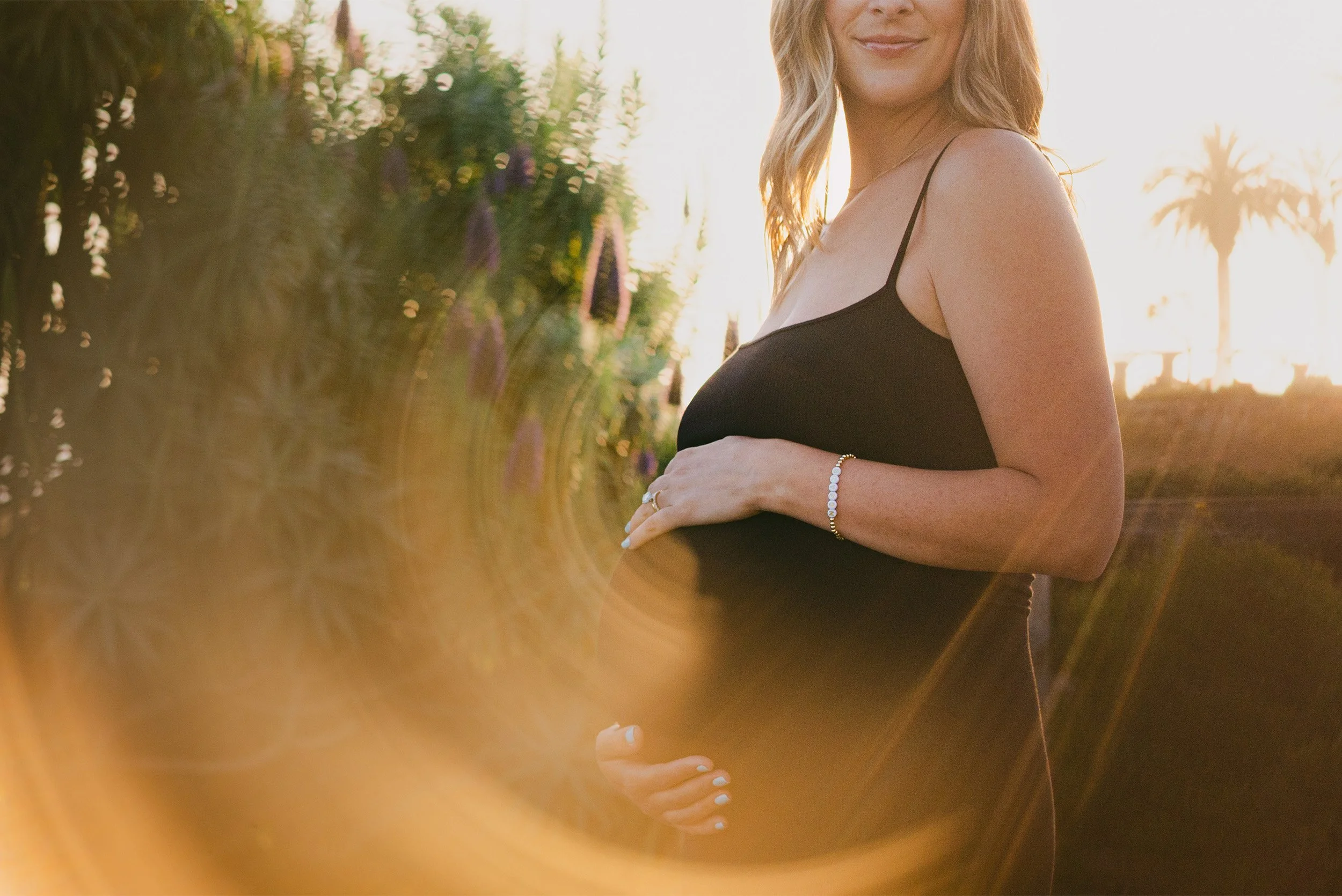 Close-up of pregnant belly in black dress with golden sunset light flare, Orange County maternity photographer