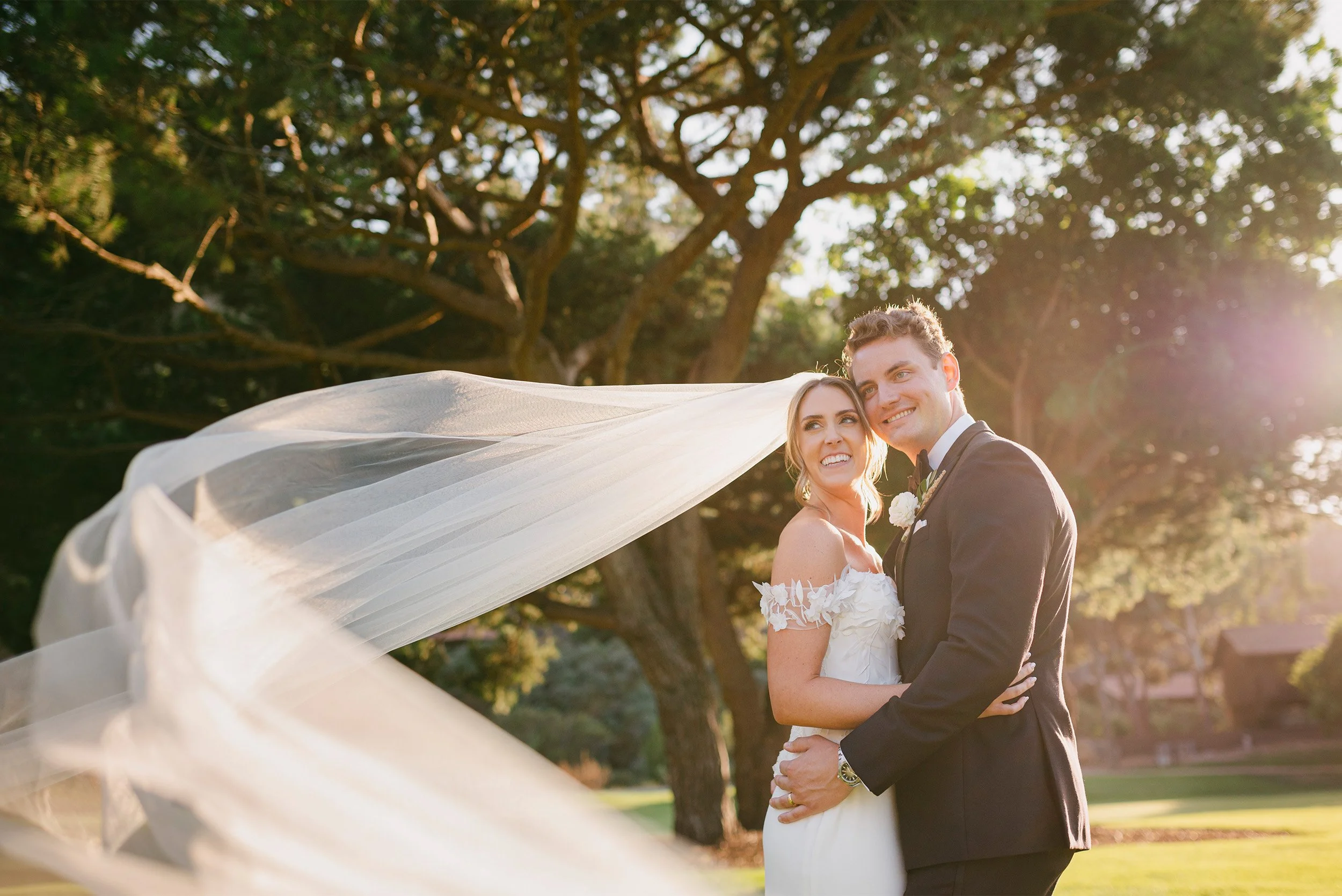 Highly recommended wedding photographer at The Ranch at Laguna Beach captures bride and groom smiling as veil dramatically blows across sunlit tree backdrop