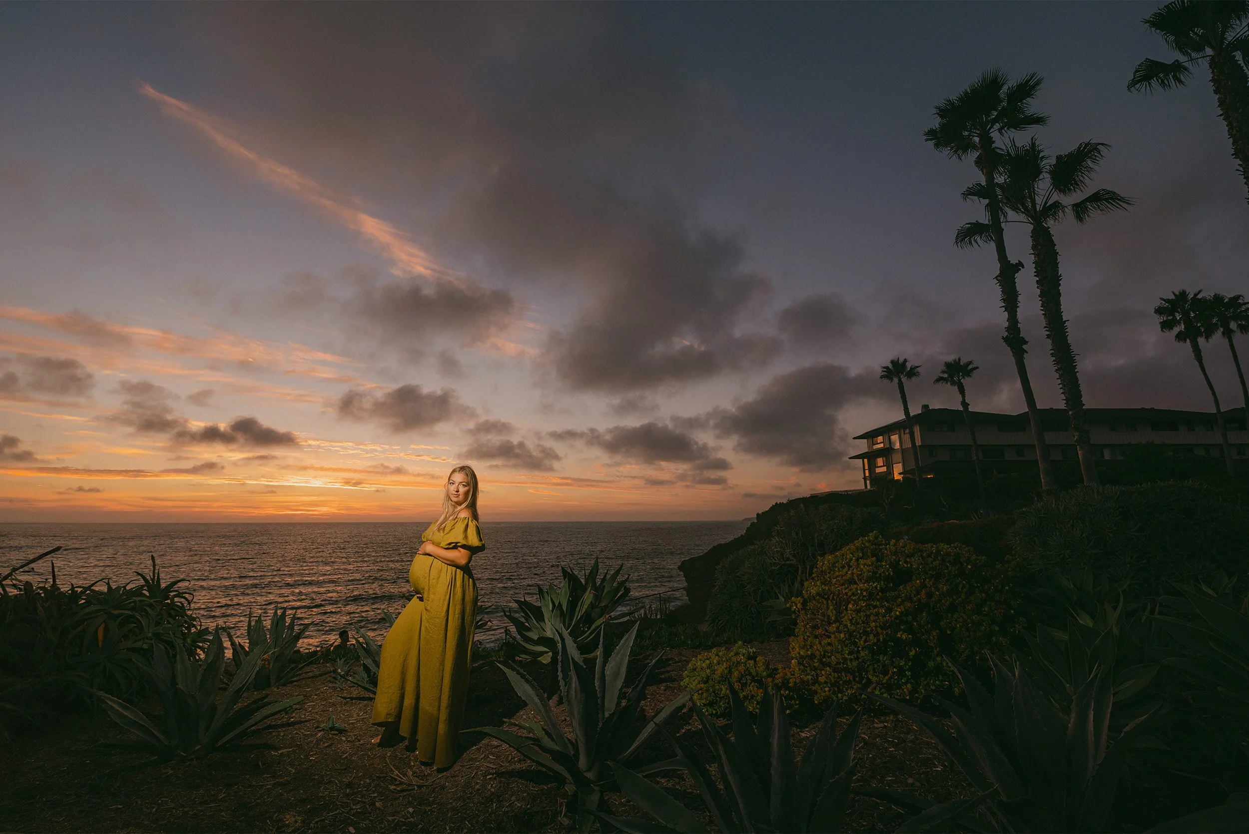 Pregnant woman in mustard dress standing on coastal bluff at dusk, award-winning maternity photographer in Orange County