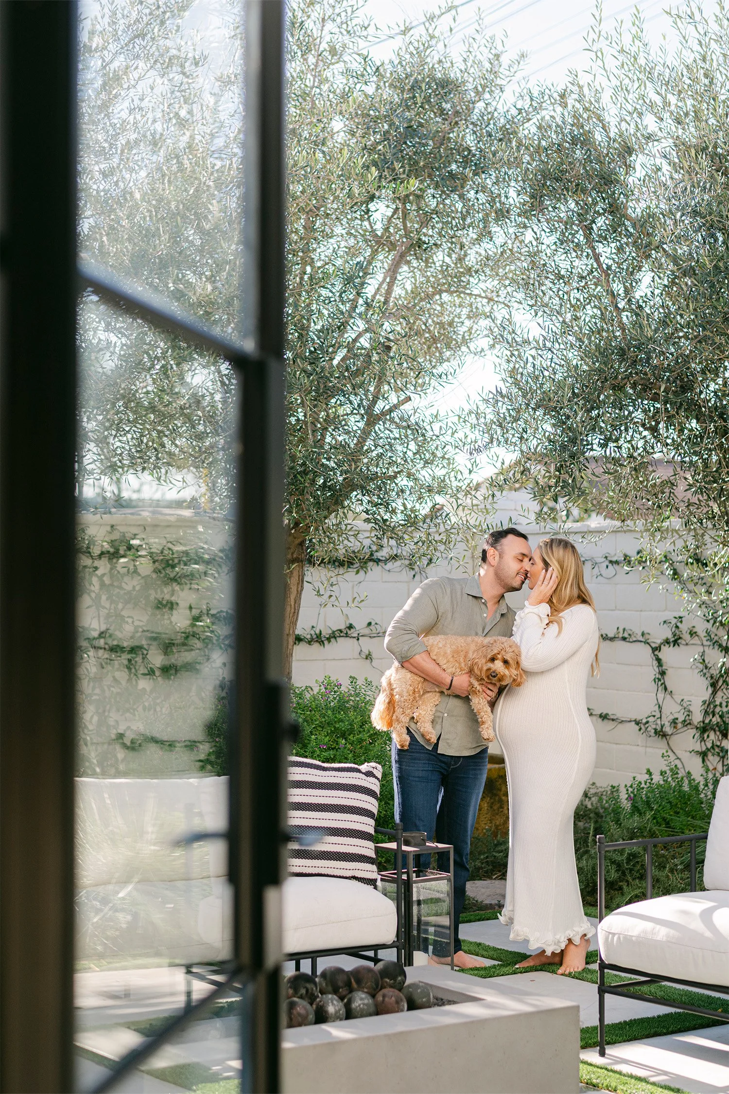 Pregnant couple kissing with dog in lush backyard garden, highly recommended maternity photographer Orange County