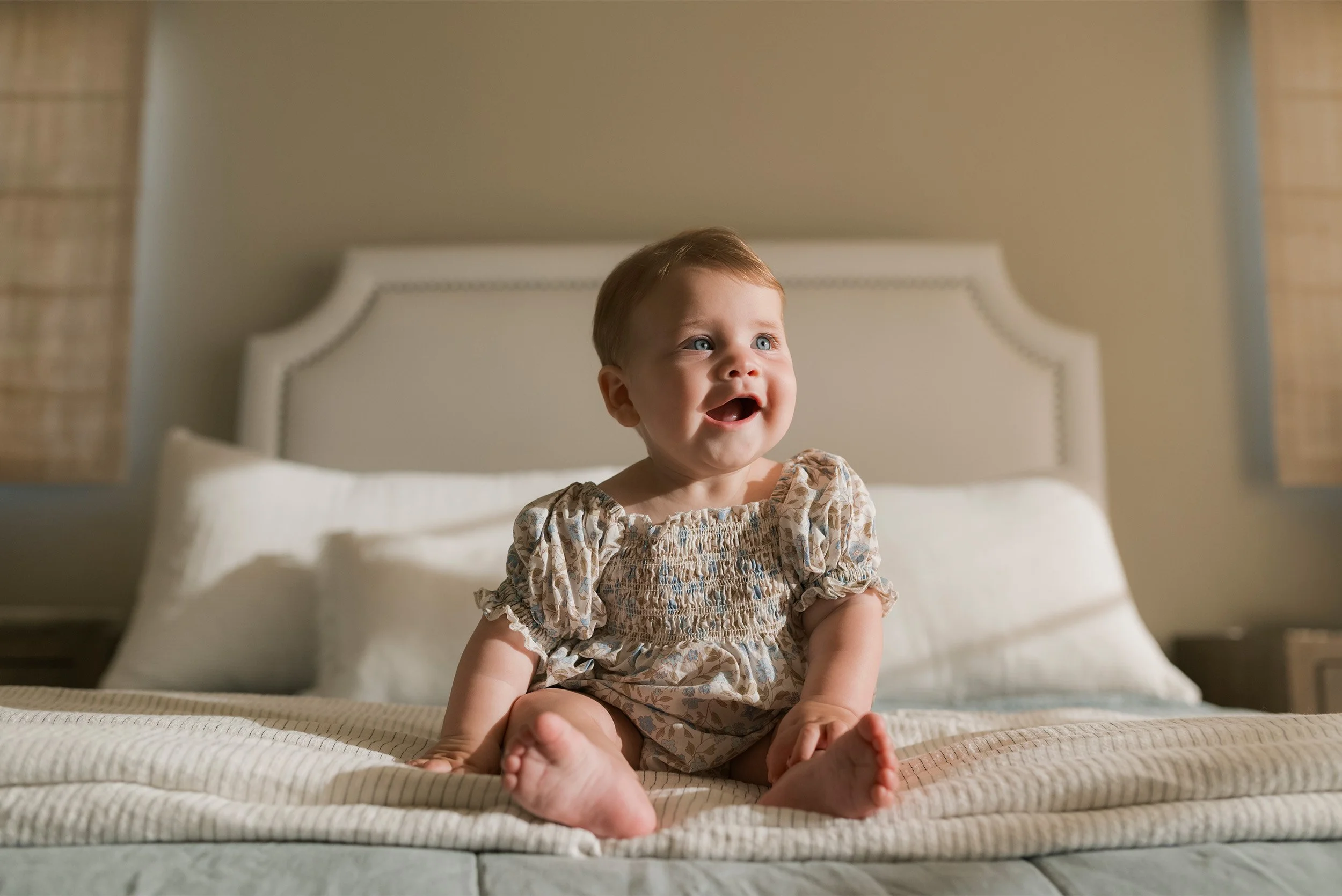 Lifestyle family photography in Orange County featuring smiling baby sitting on bed in natural light