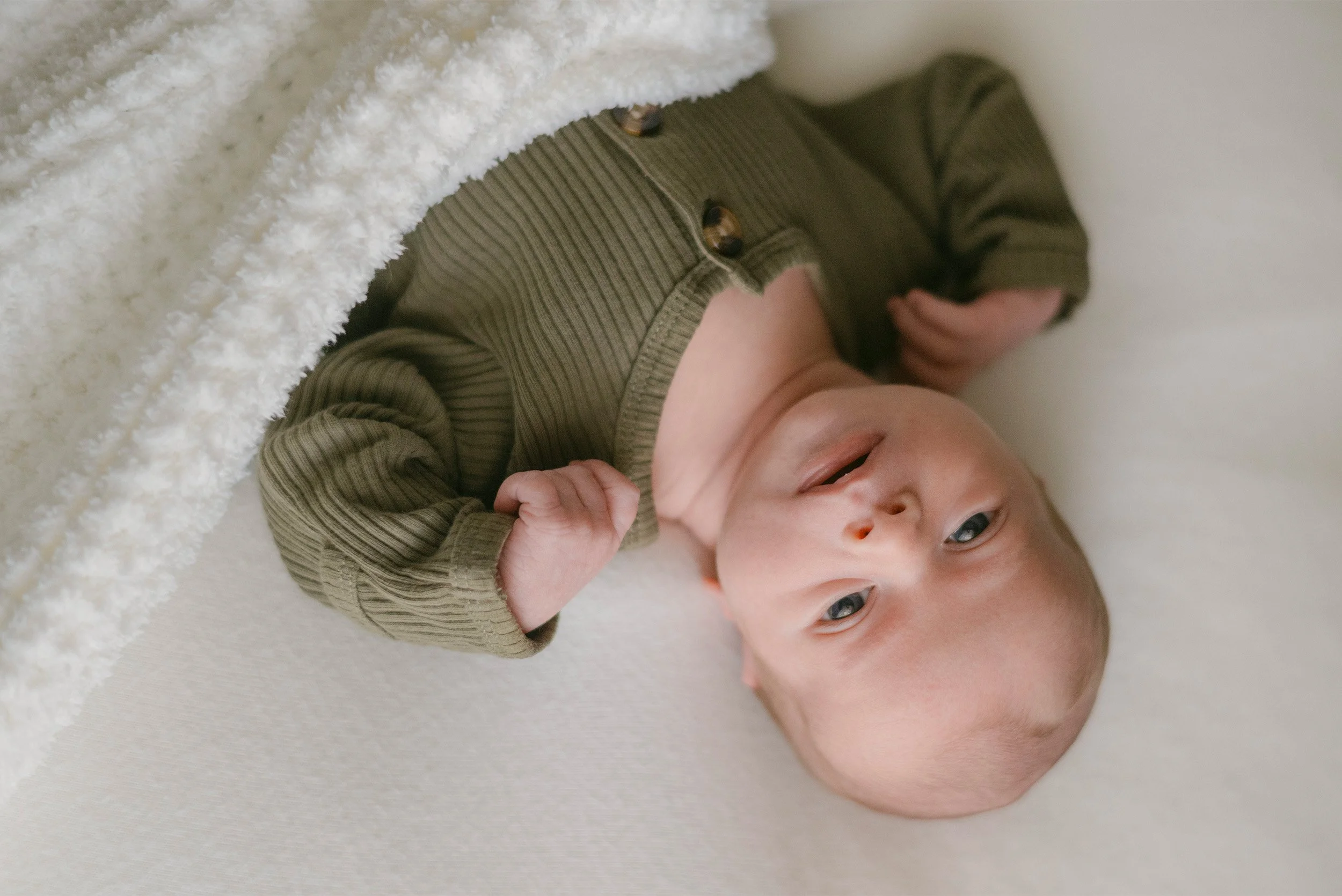 Close-up of smiling baby in olive knit outfit peeking from blanket, highly recommended baby photographer Orange County