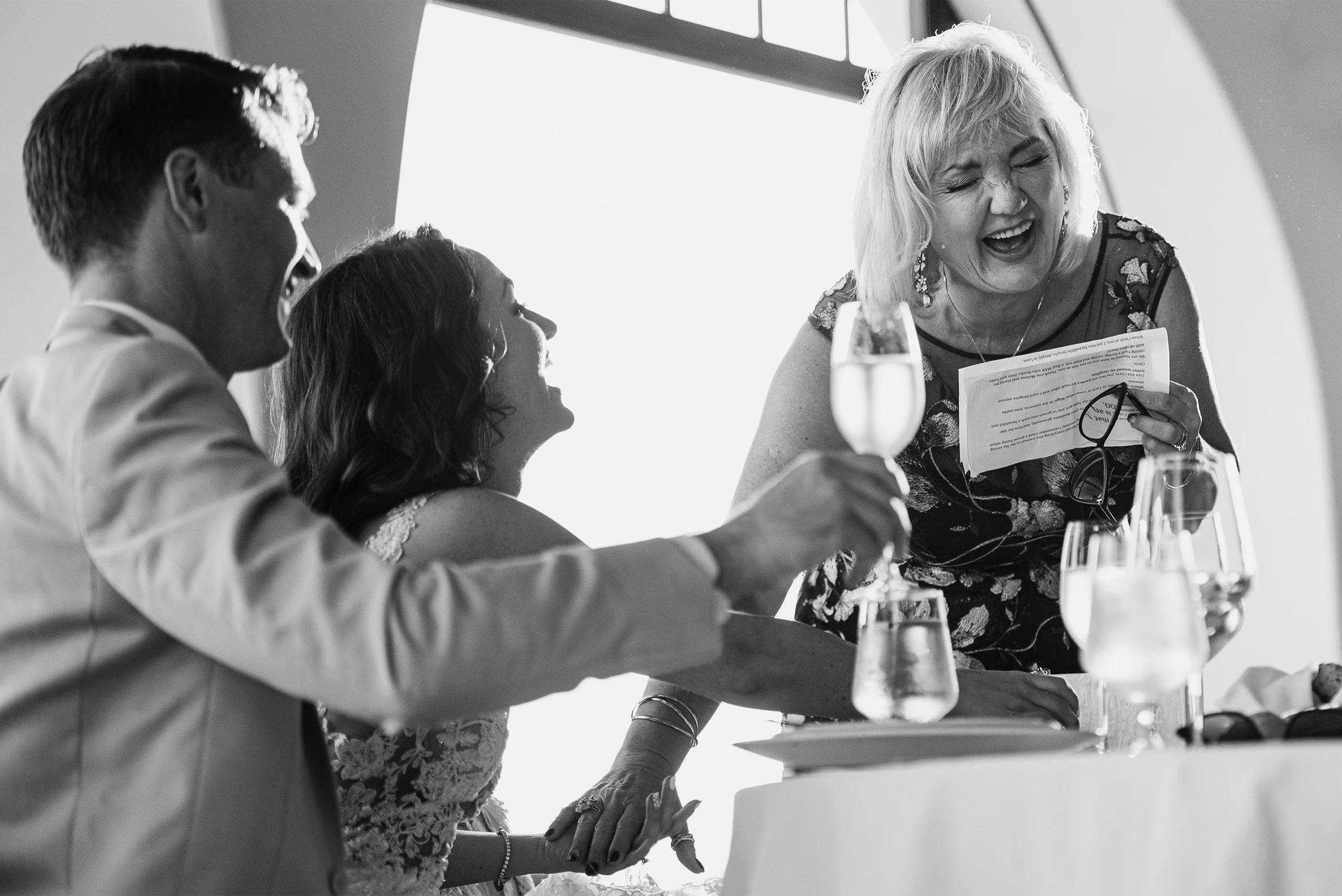 Santa Barbara wedding photographer captures candid laughter between bride, groom, and guest at reception table, black and white