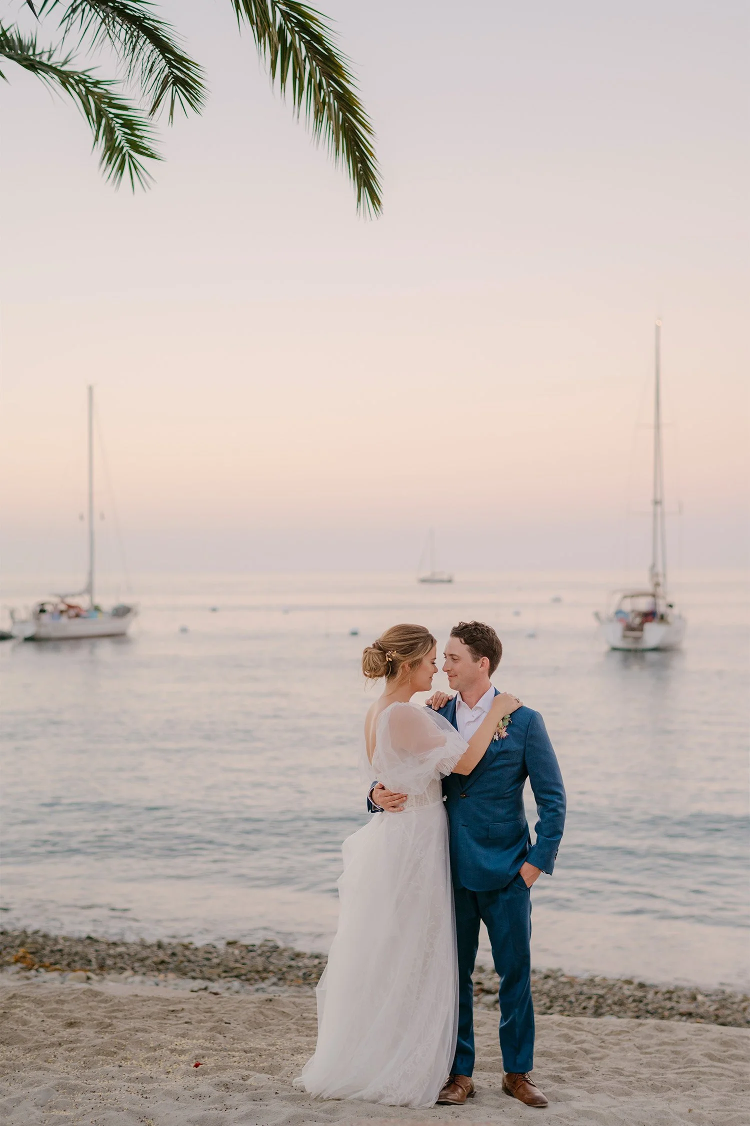 Bride and groom embracing on Catalina Island beach at sunset with sailboats in background, coastal wedding photography