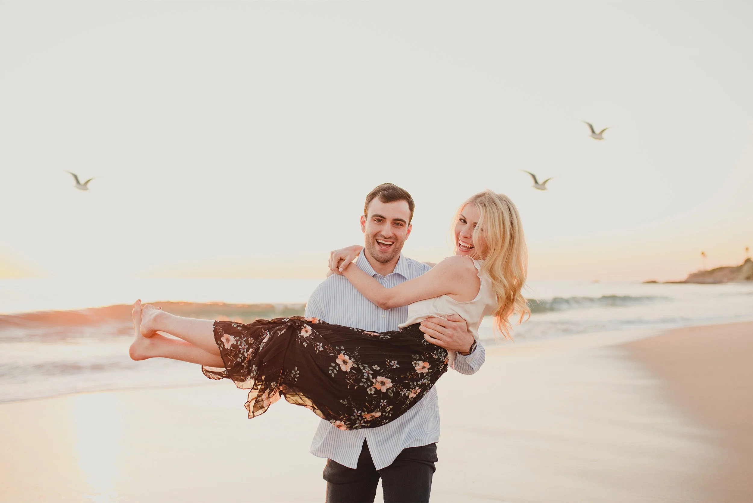 Groom-to-be carrying laughing bride-to-be on the beach with seagulls flying overhead, coastal laguna beach engagement photography