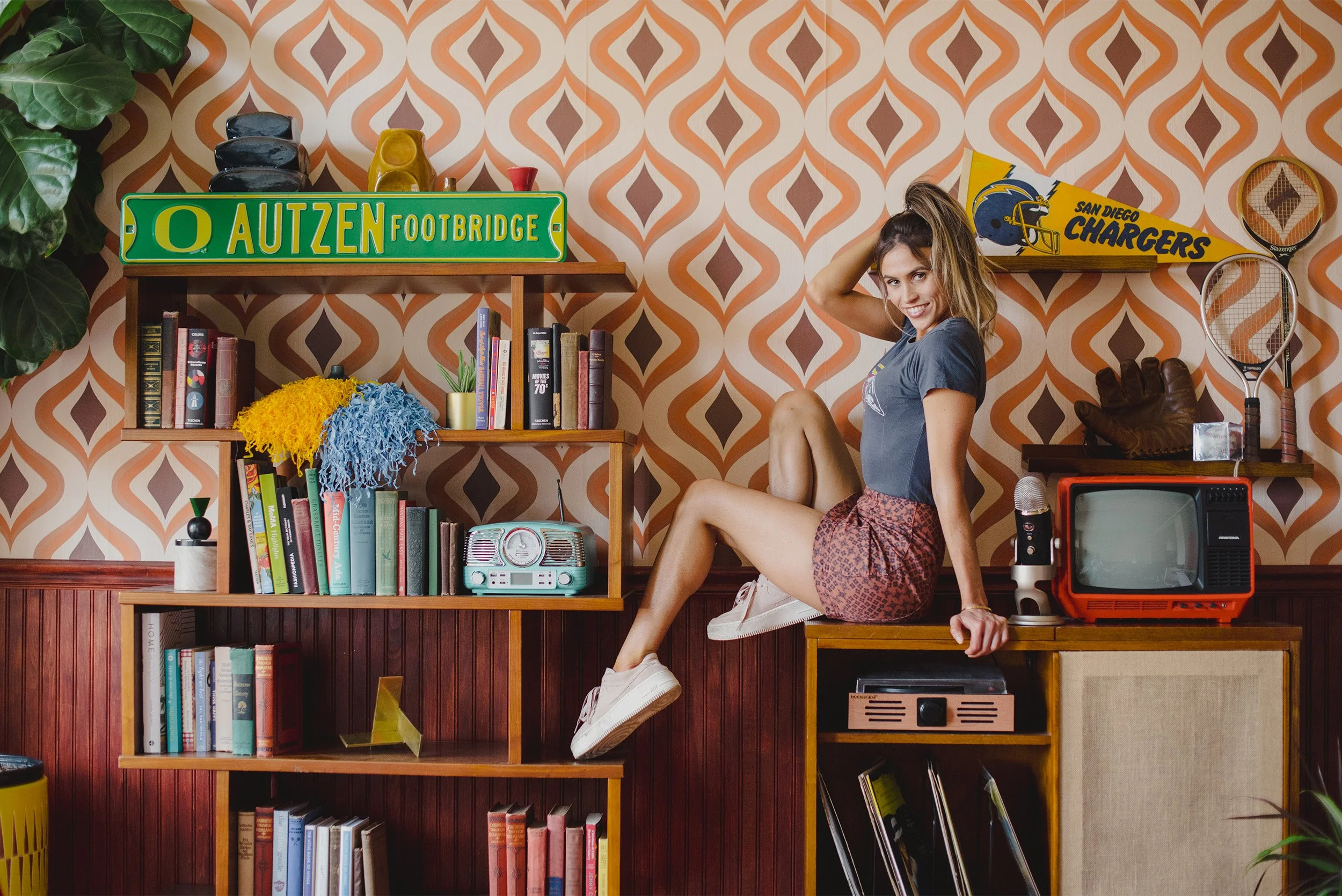 Orange County lifestyle photographer captures woman perched on vintage record cabinet surrounded by retro decor and sports memorabilia
