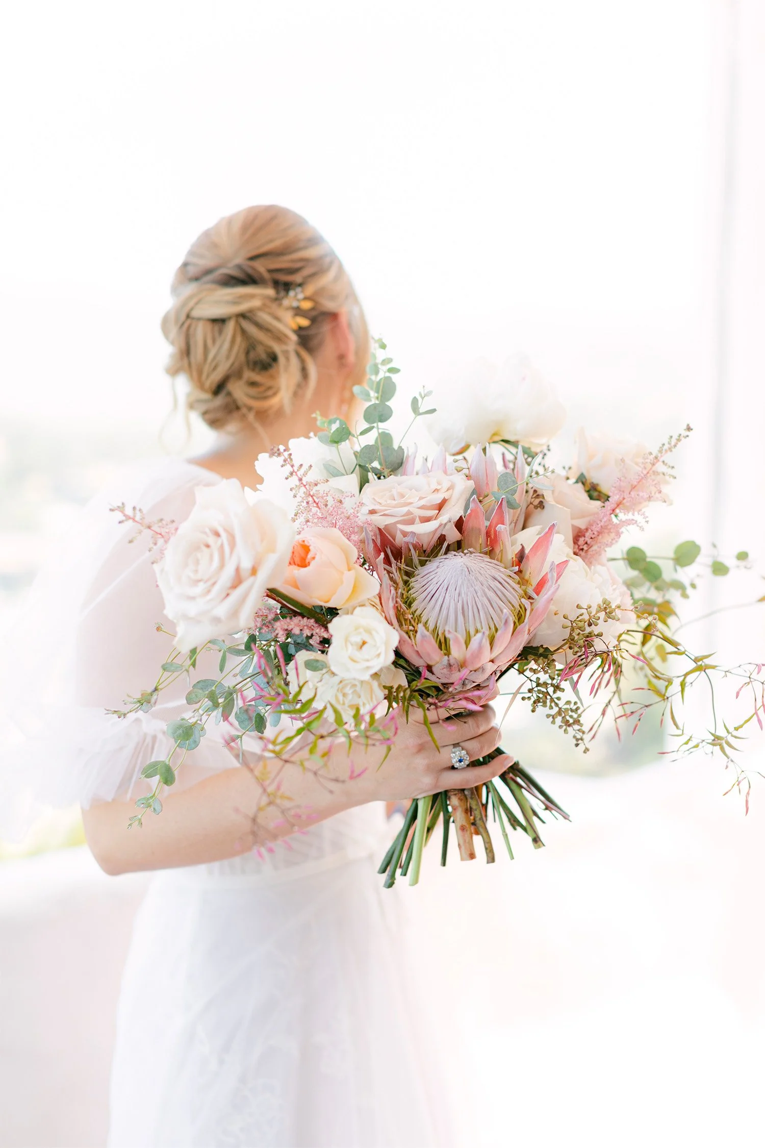 Bride from behind holding lush protea and rose bouquet, soft bridal portrait